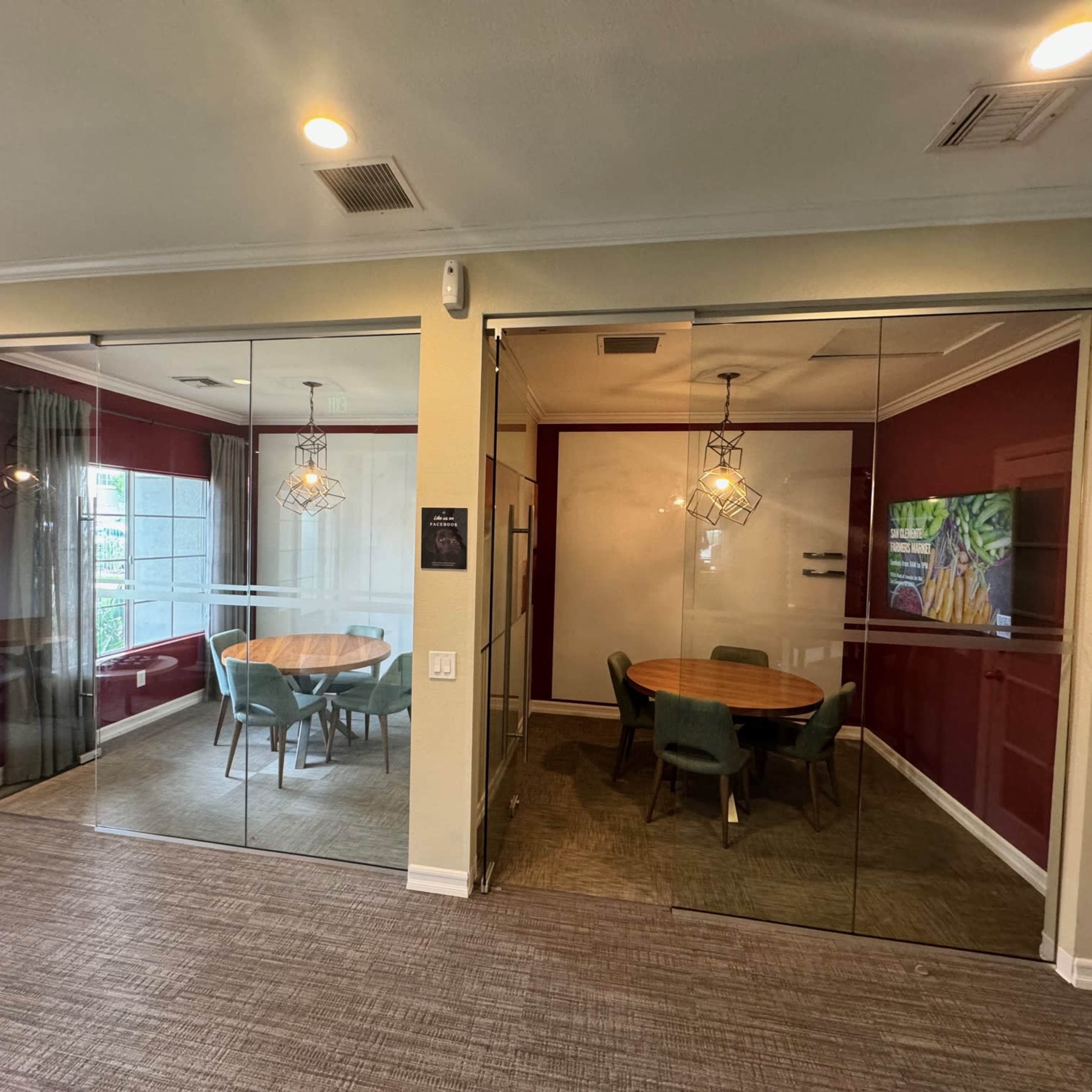 A modern office space with two glass-walled conference rooms, one featuring a round wooden table and green chairs, and the other a seating area with a view outside.