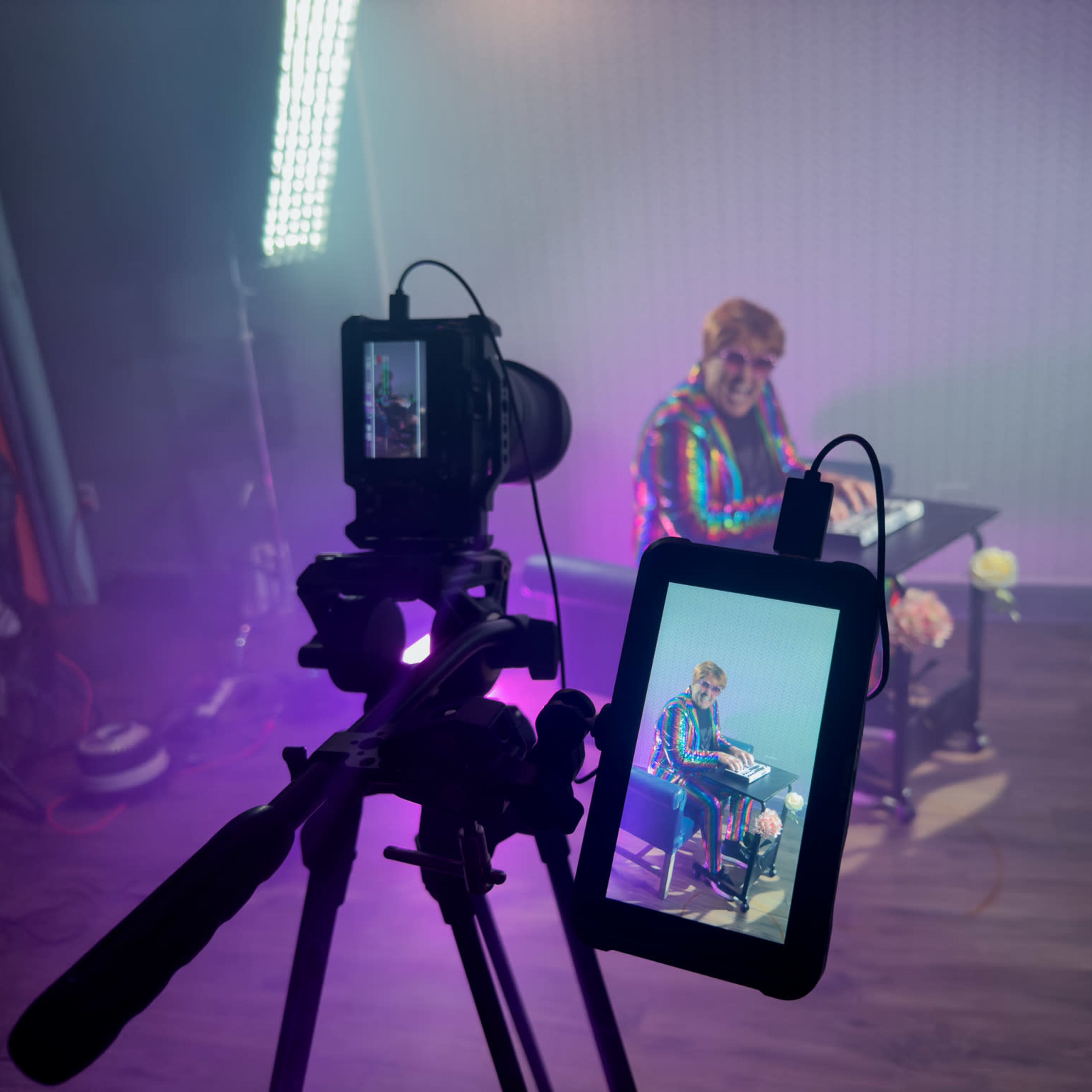 A person is sitting at a desk under colorful lighting, while a camera on a tripod captures the scene on a monitor.
