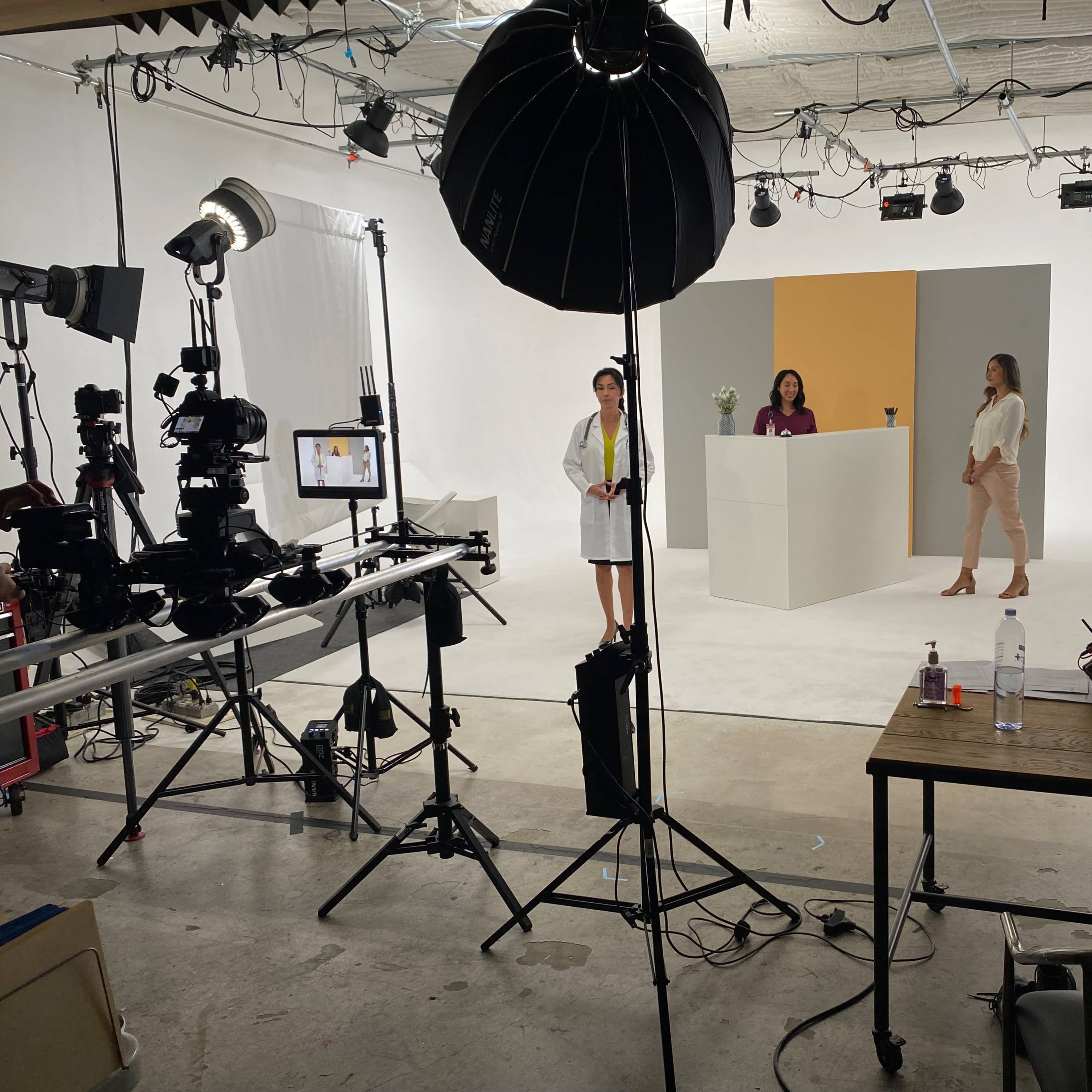 A studio setup with two women standing in front of a white podium, surrounded by lighting equipment and cameras, while additional screens display images.