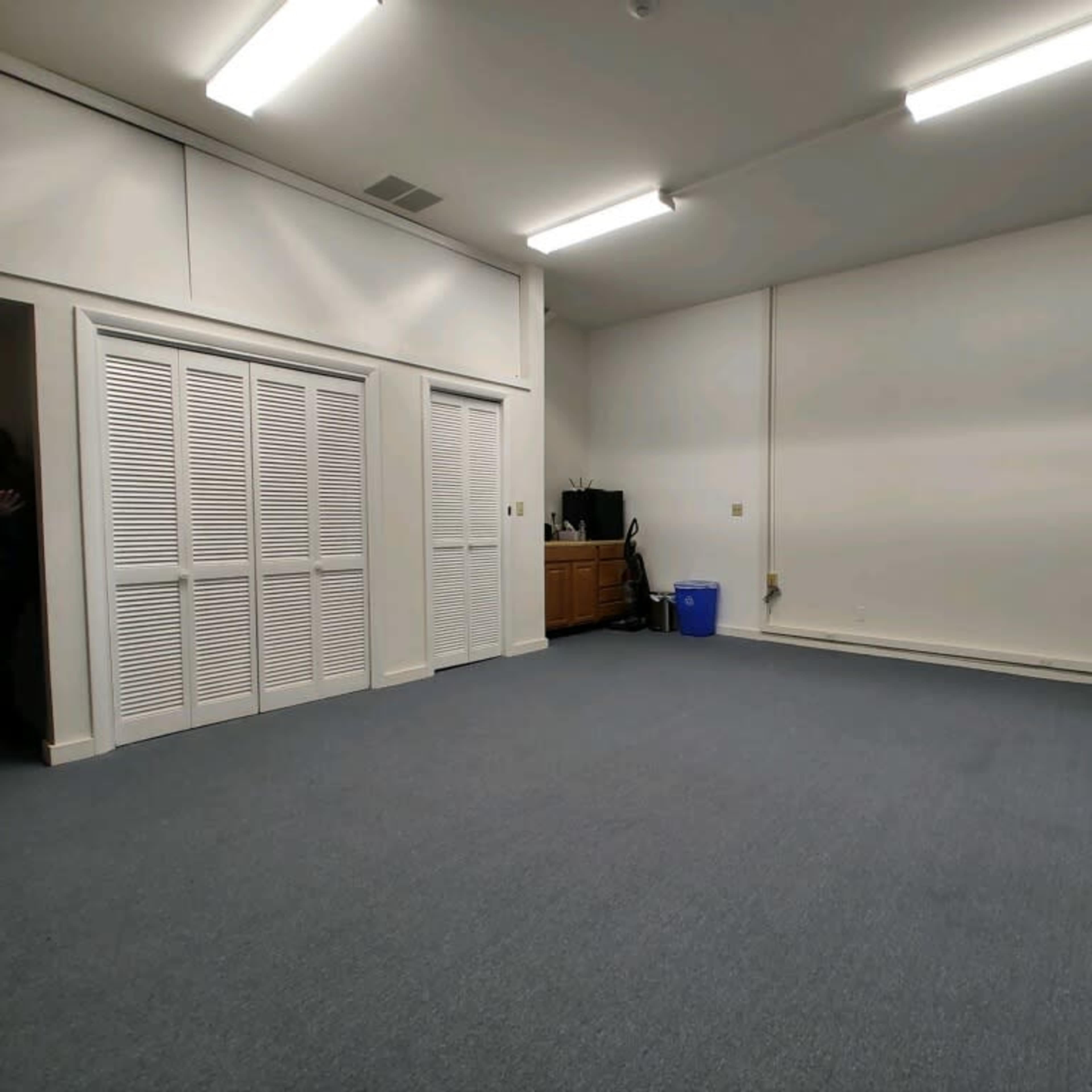A spacious, empty room with blue carpet, white walls, and minimal furniture including a yellow stool and a blue container.