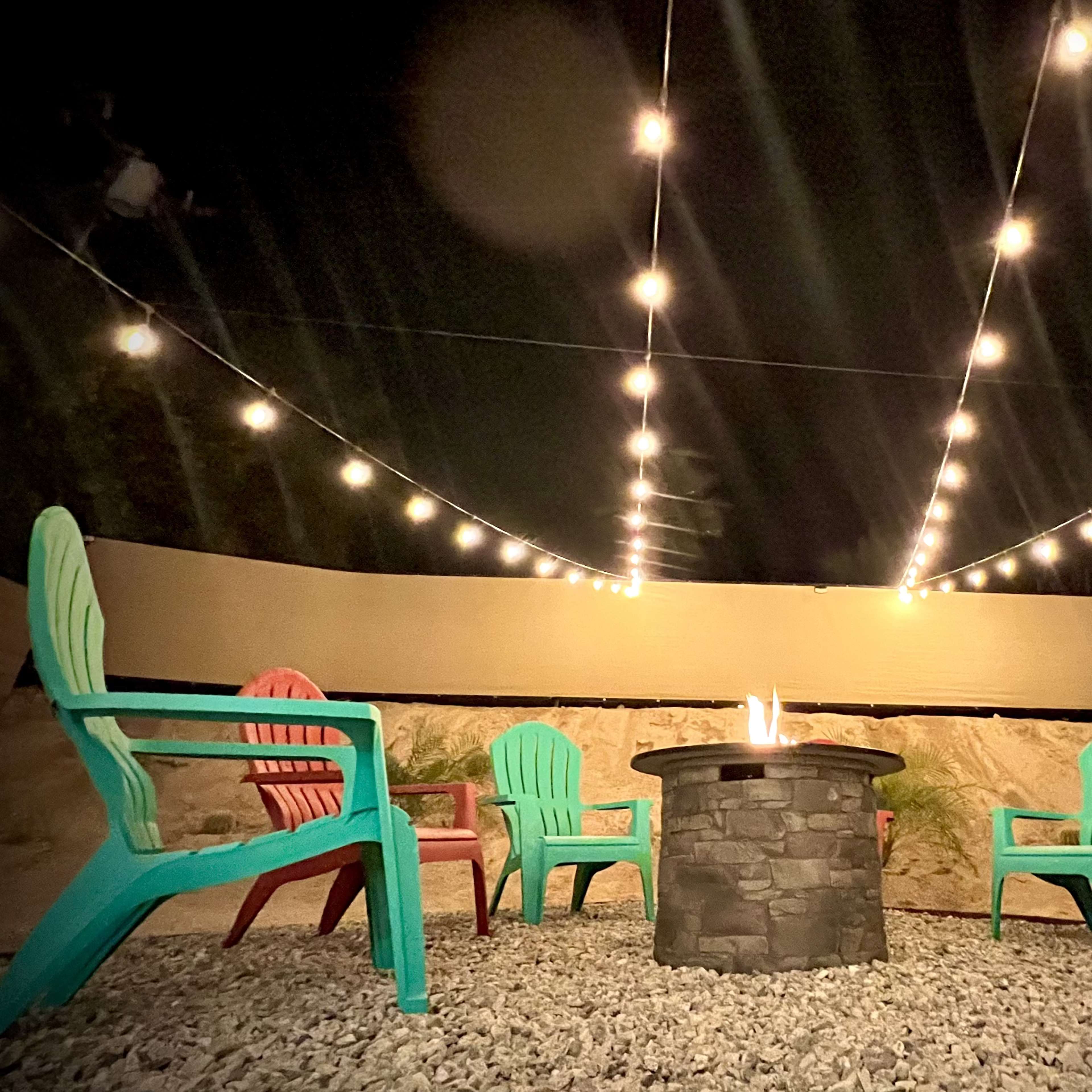 A circular stone fire pit is surrounded by colorful adirondack chairs under a string of lights at night.