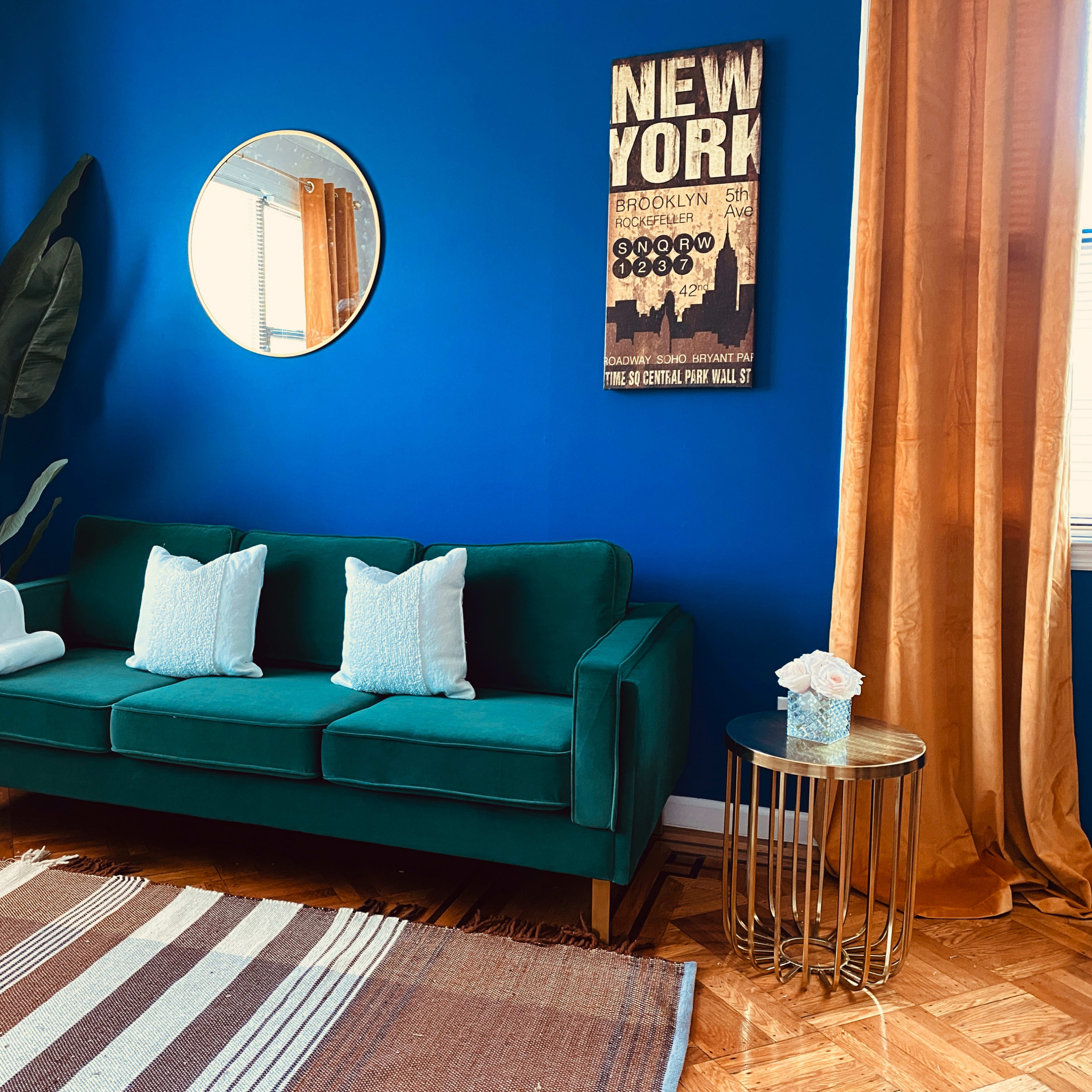 A green sofa with decorative pillows sits against a blue wall, accompanied by a mirror and a plant, with golden curtains framing a window and a layered rug on the floor.