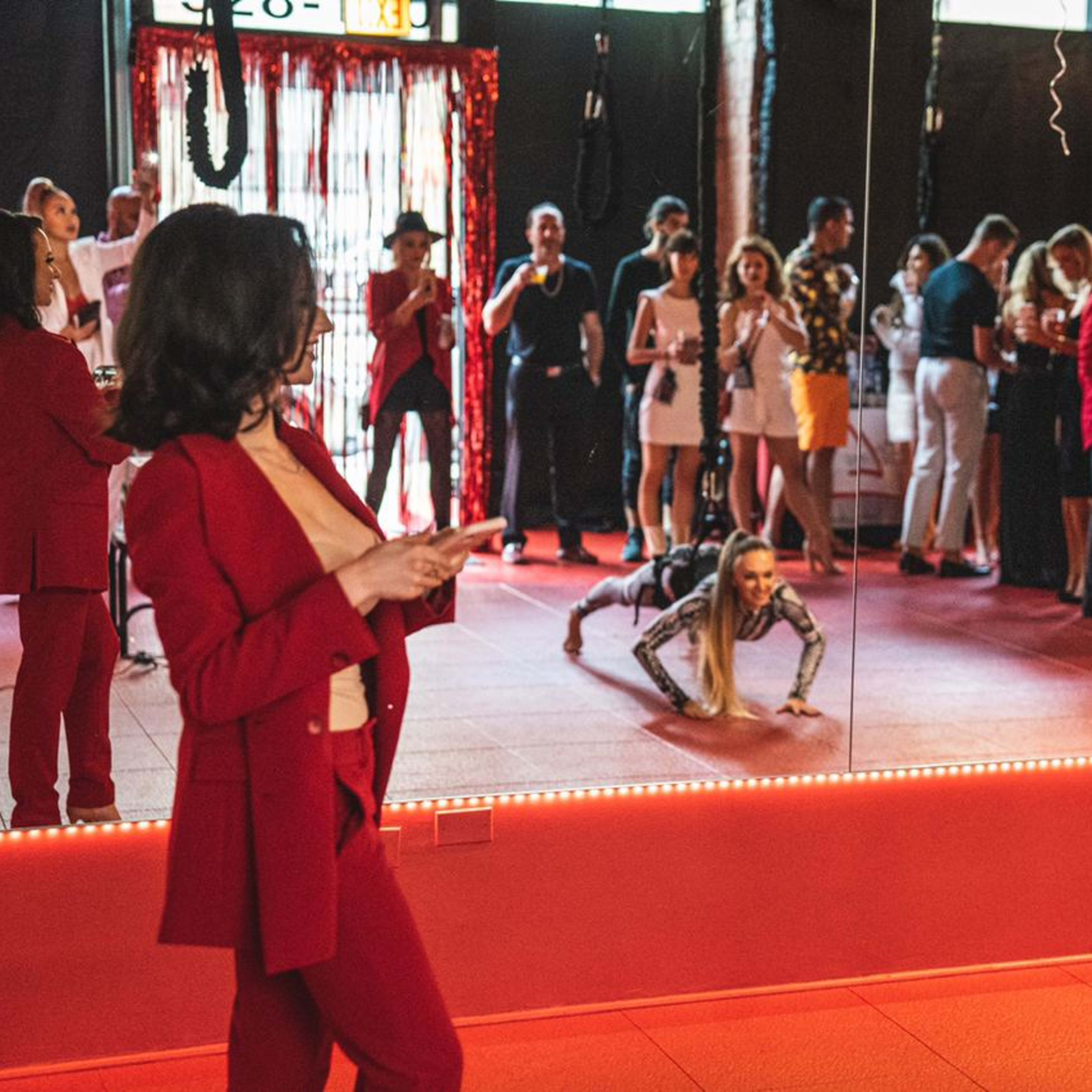 A group of people socialize in a mirrored room, while a woman in a red suit checks her phone and another performs push-ups.