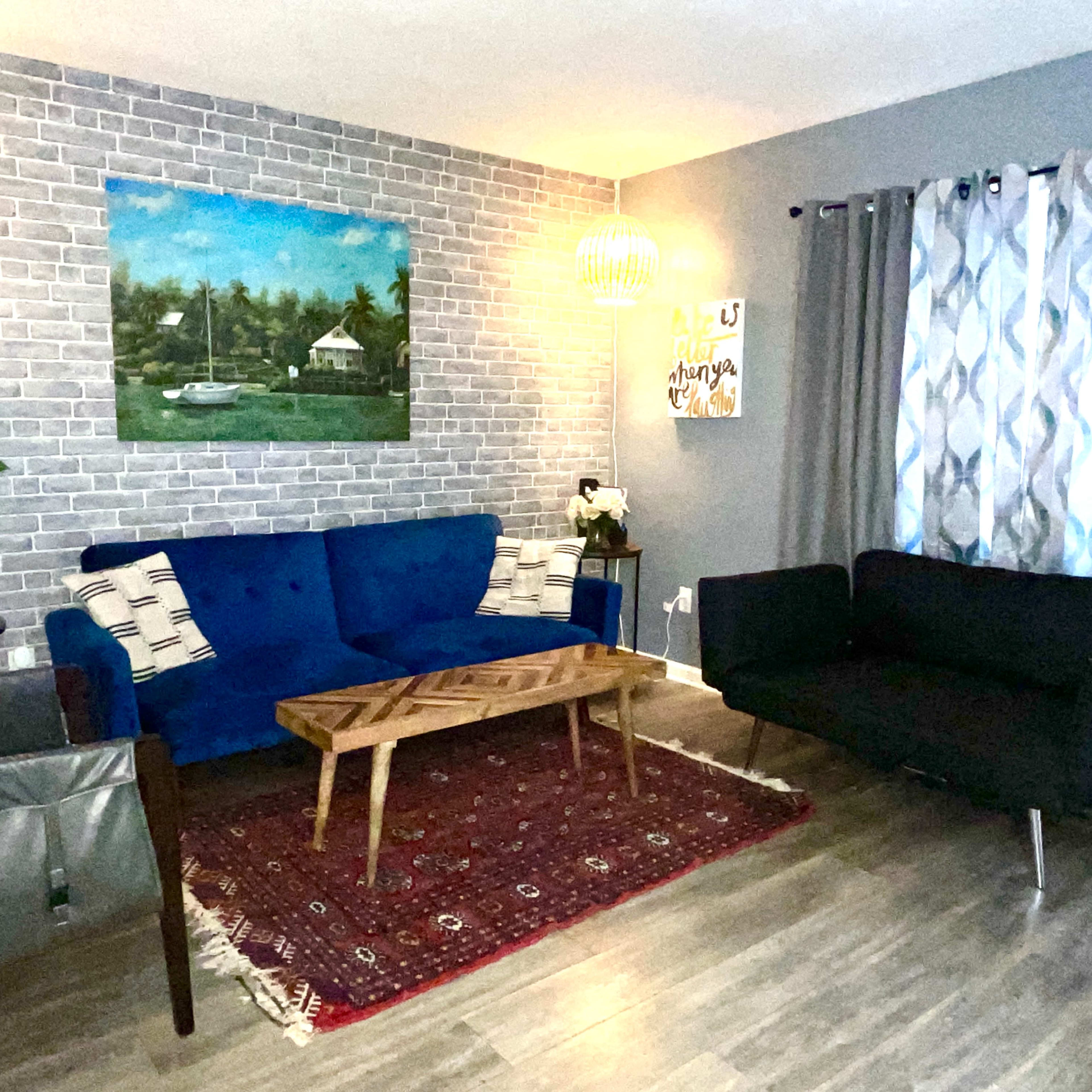 A living room with a blue sofa, a black loveseat, a wooden coffee table, and a large wall painting, all arranged on a hardwood floor with an area rug.