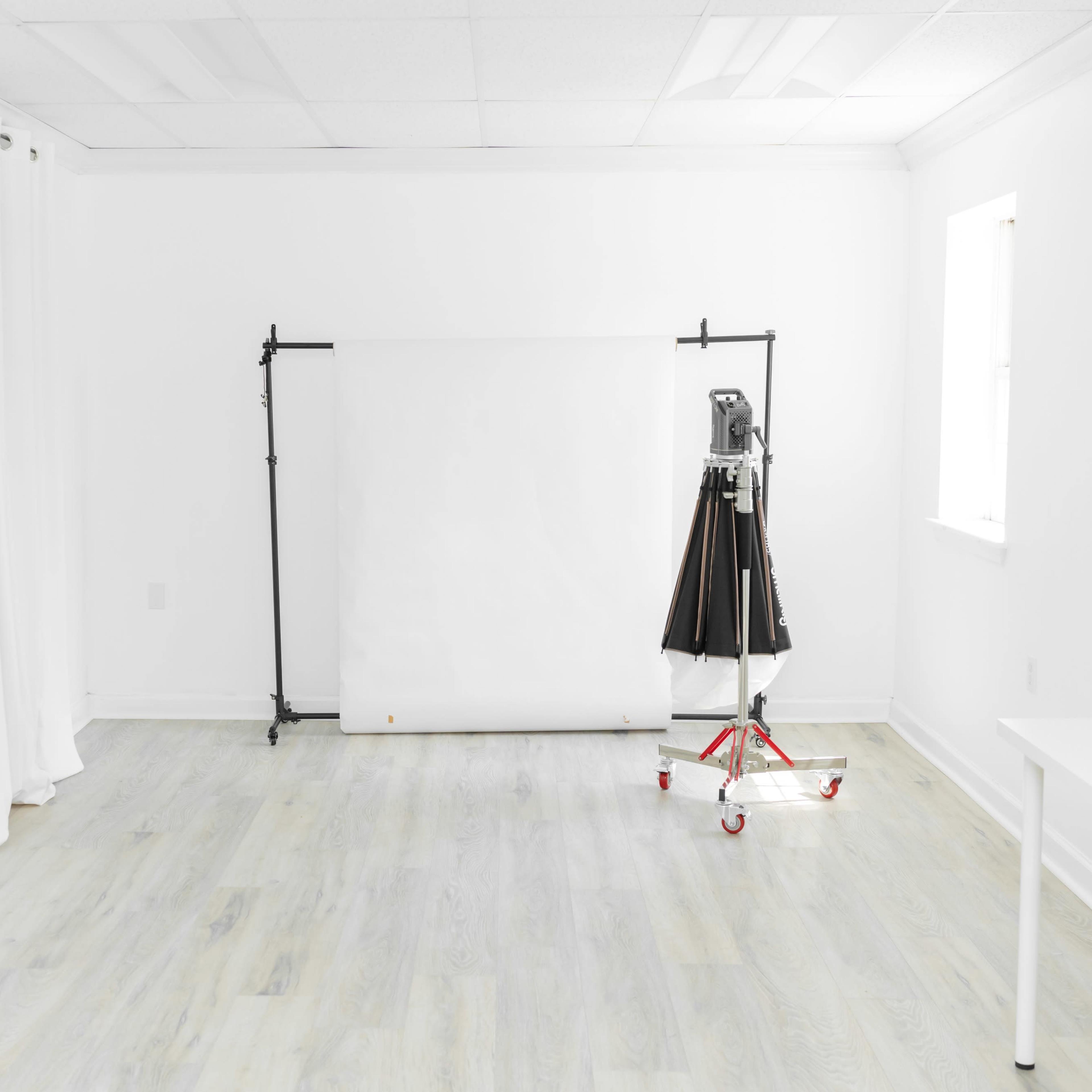 A bright, minimalist studio features a blank white backdrop, a lighting rig on wheels, and white curtains along one wall.
