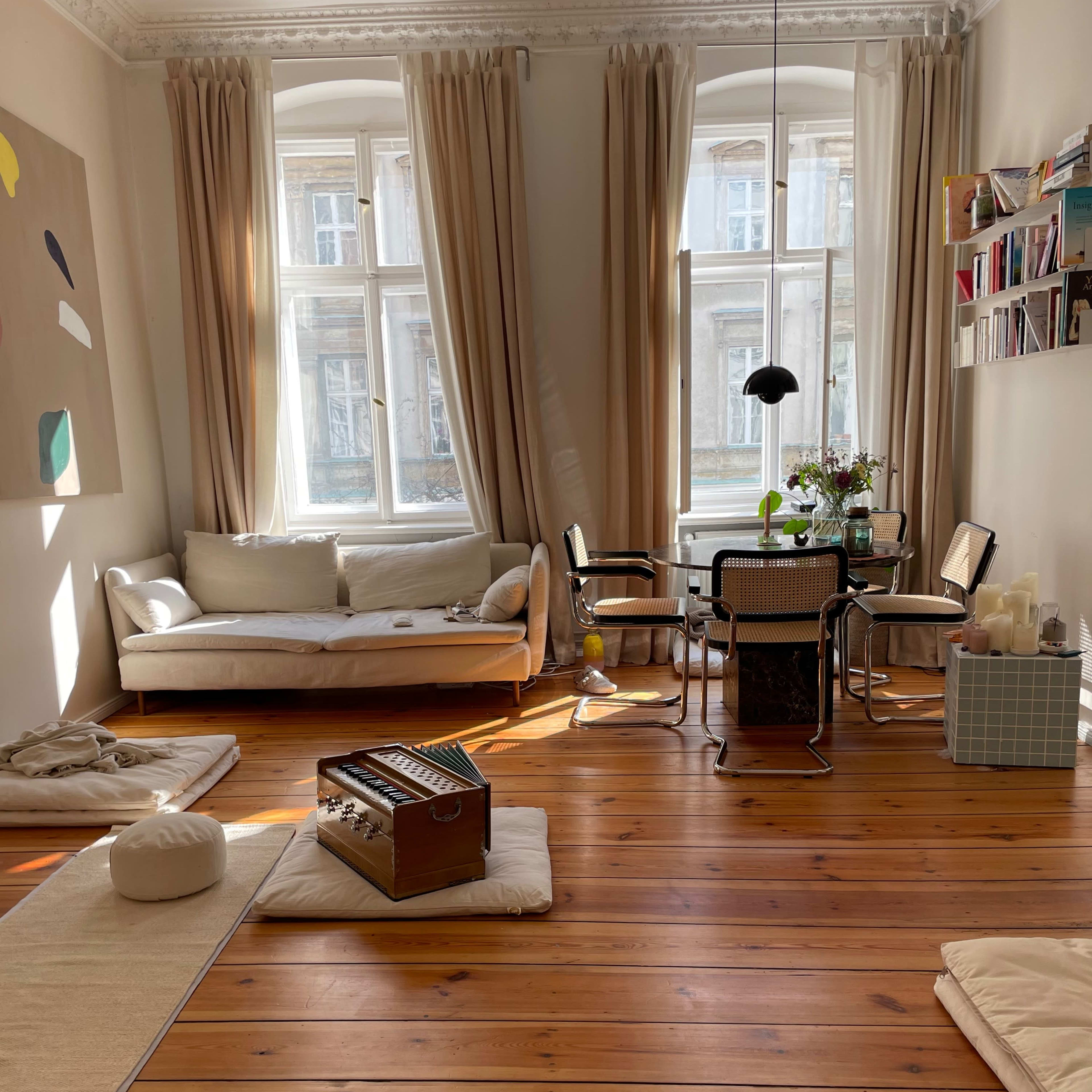 The room features a light-colored sofa, a dining table with chairs, and wooden flooring, illuminated by natural light coming through the large windows.