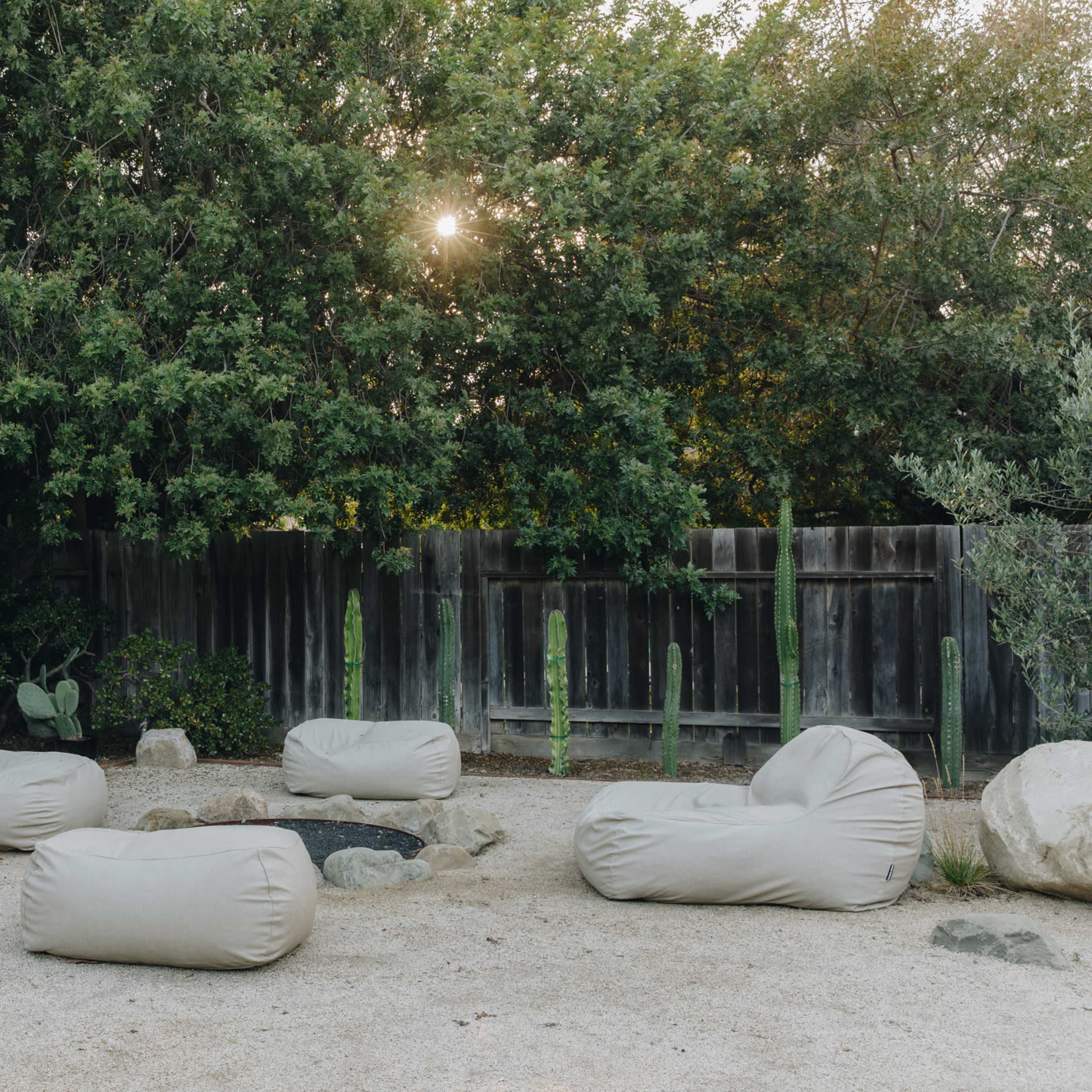 A gravel area features several beige bean bag chairs arranged around a fire pit, with a black shed and wooden fence in the background.