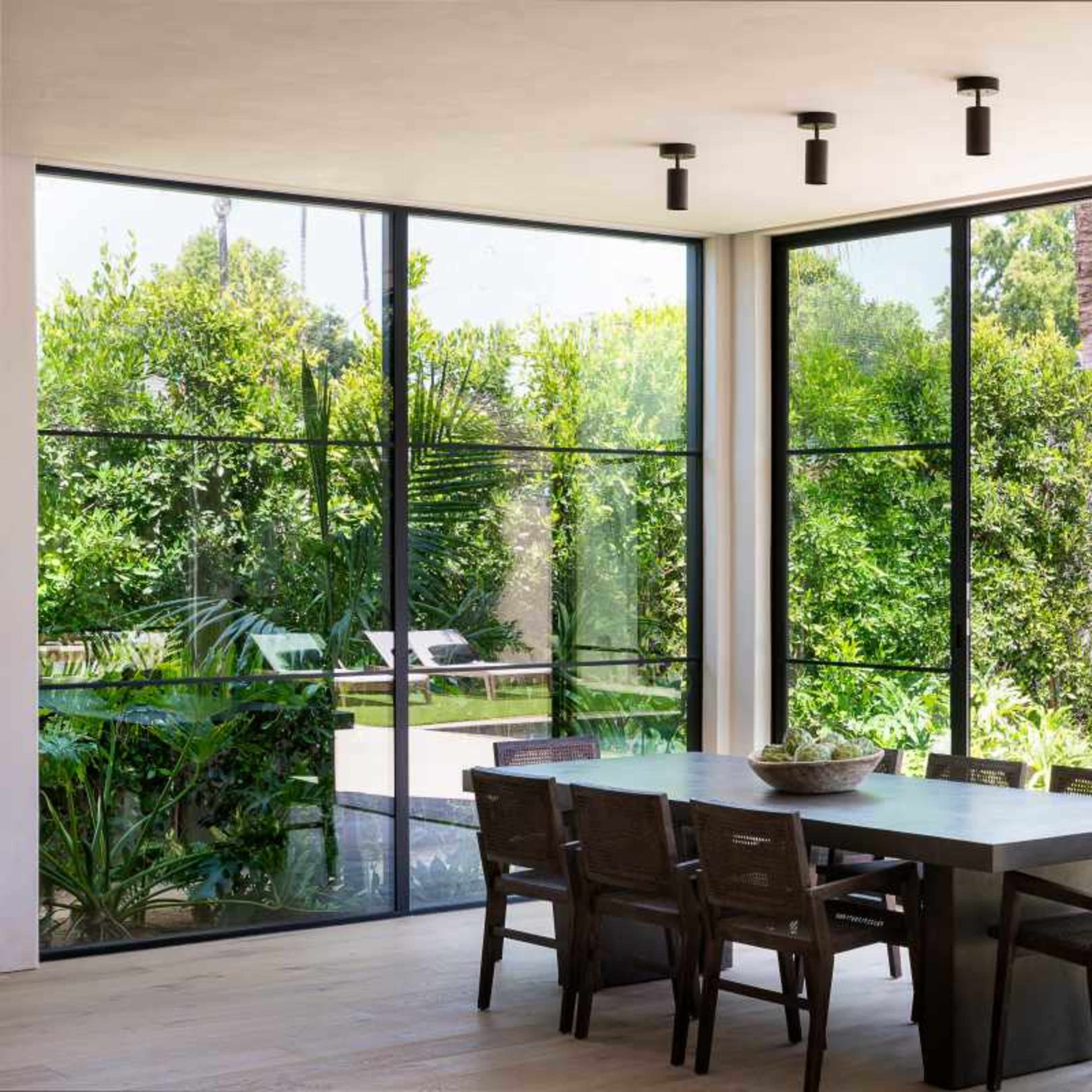 A modern dining area with a long table and chairs, surrounded by large windows that open to a green outdoor space featuring palm trees and an outdoor fireplace.