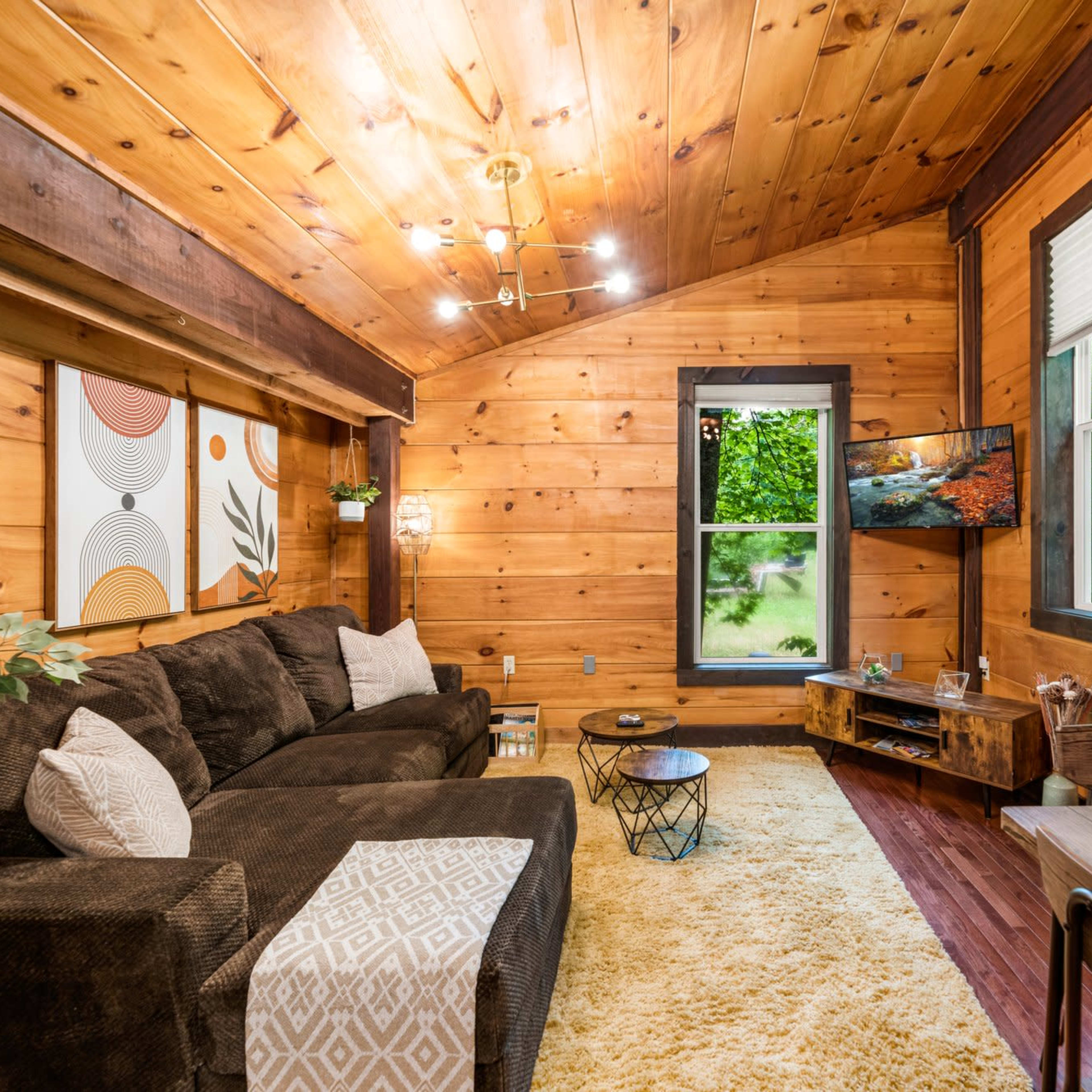 A cozy wooden room featuring a dark sectional sofa, a coffee table, and a television mounted on the wall, with large windows allowing natural light to enter.