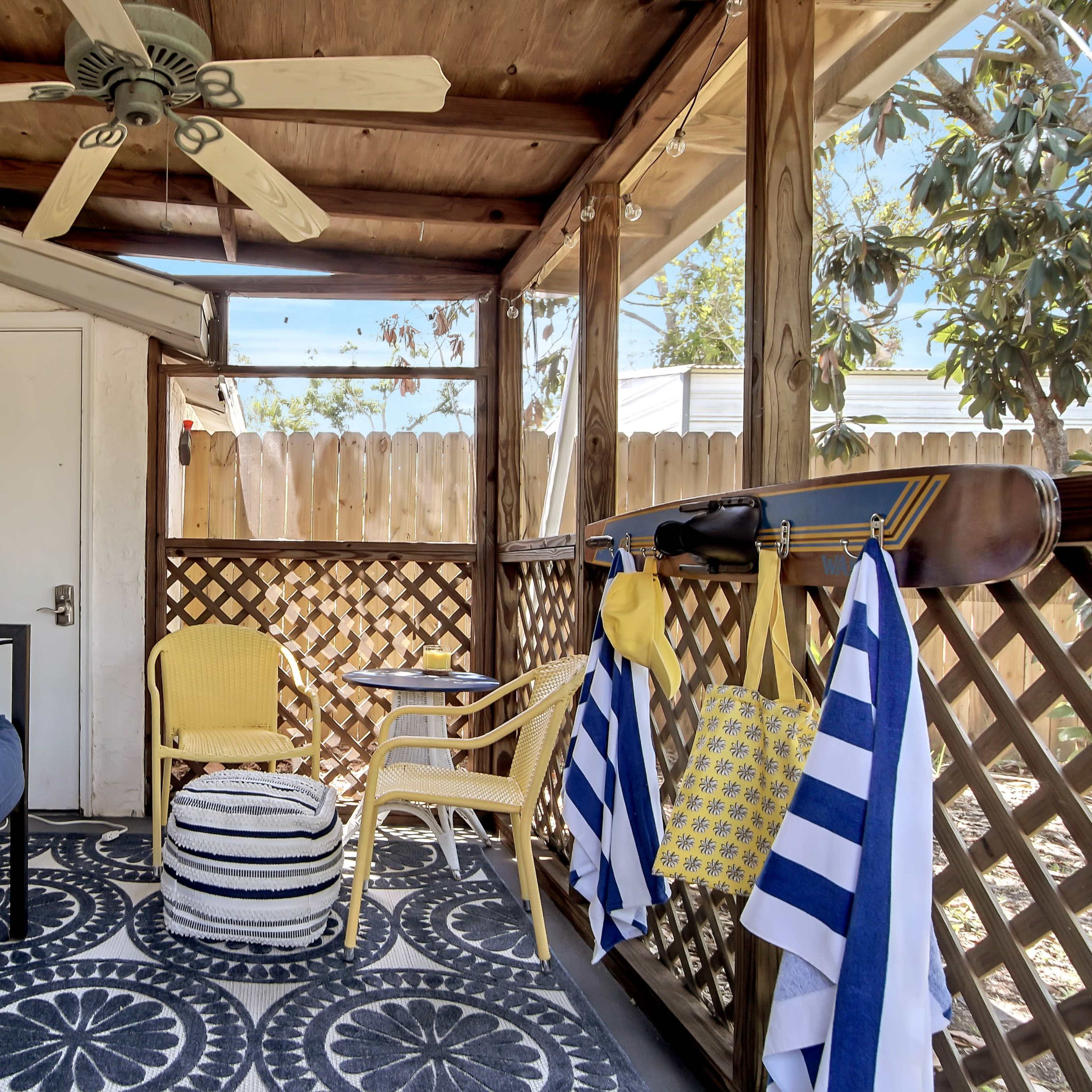 A screened porch with a blue couch, a small round table, and yellow chairs, surrounded by wooden lattice and hanging beach towels.