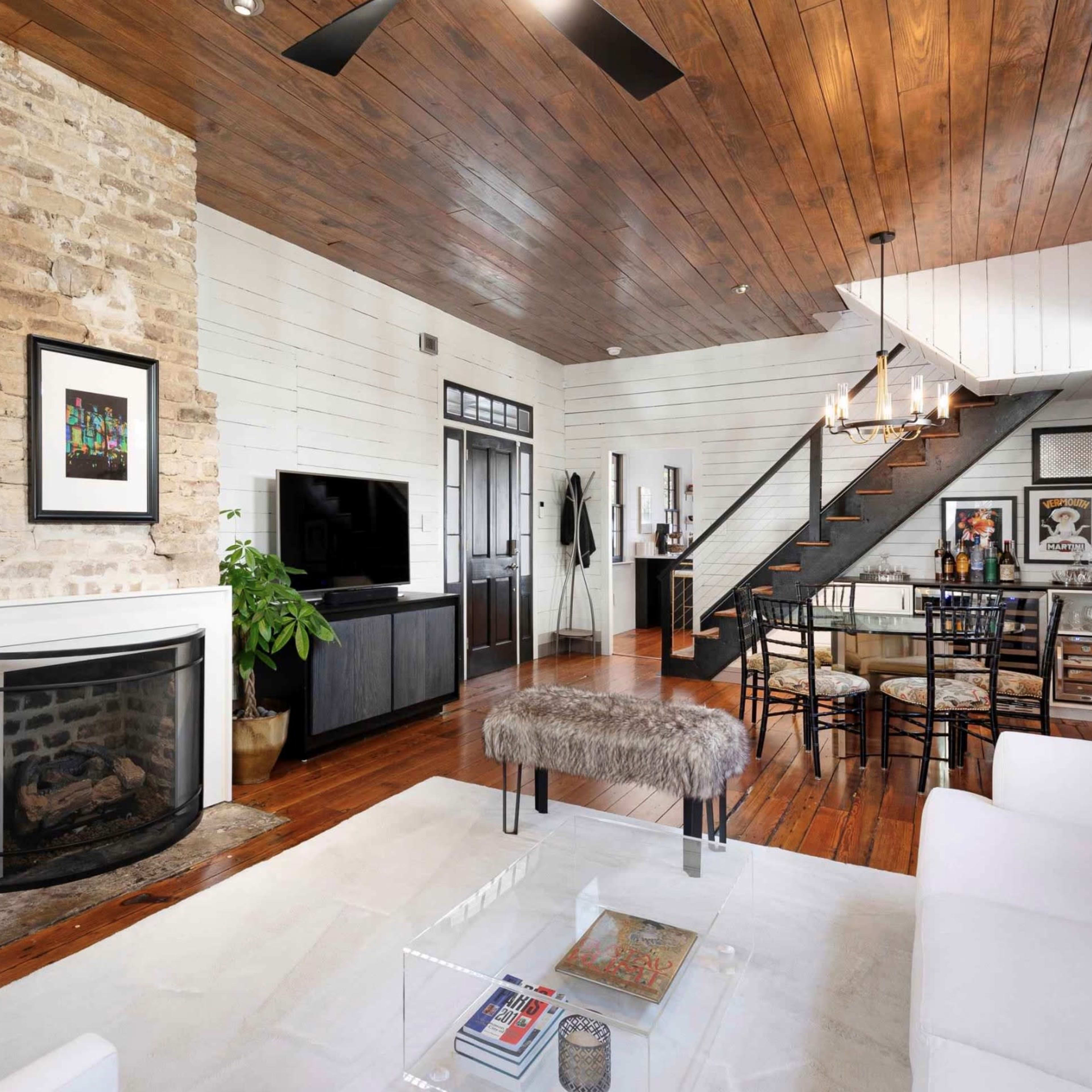A modern living area with a stone fireplace, wooden ceiling, and open staircase leading to a loft space, furnished with a white sofa and a dining table.