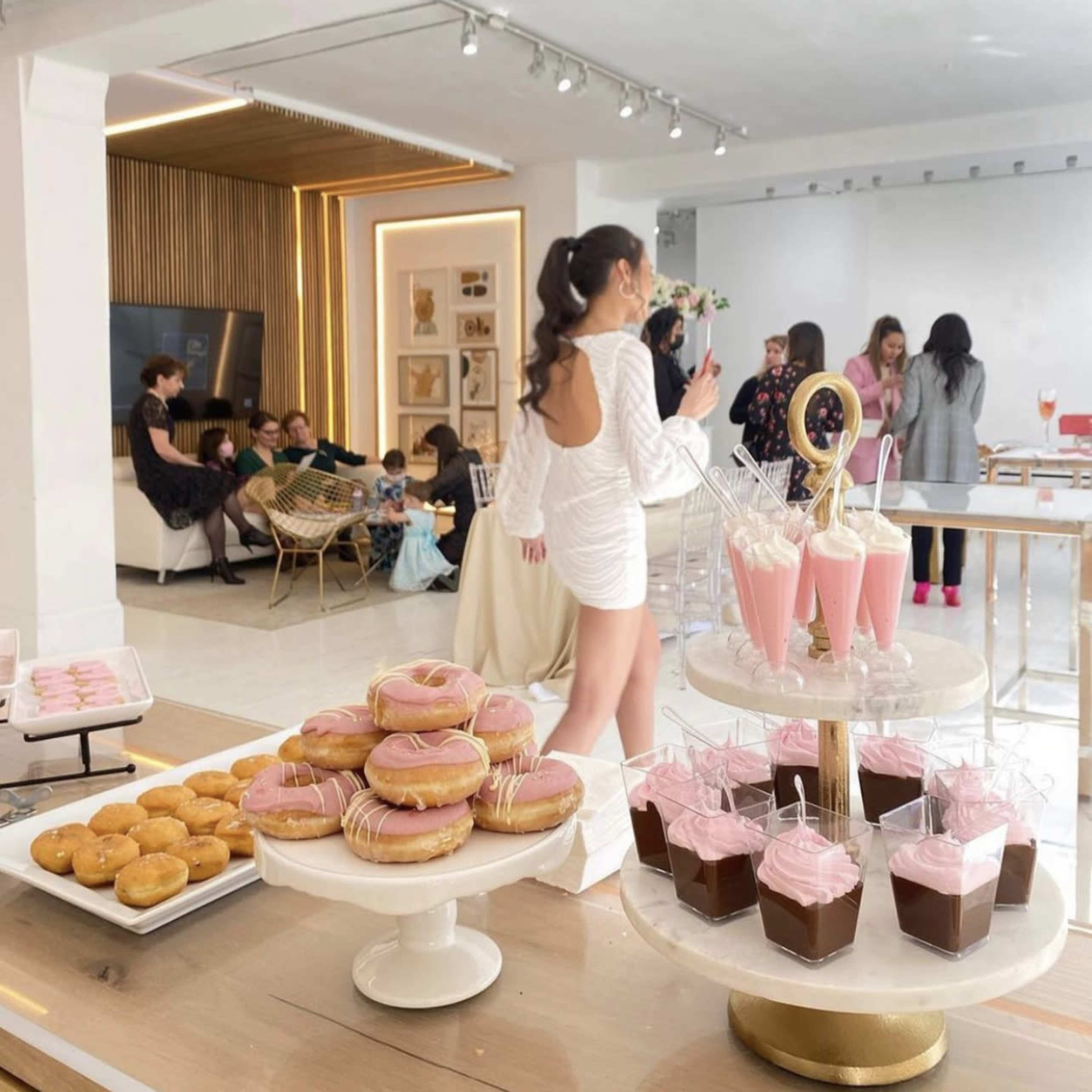 A bright room features a dessert table with assorted pastries, including donuts and pink drinks, while people socialize in the background.