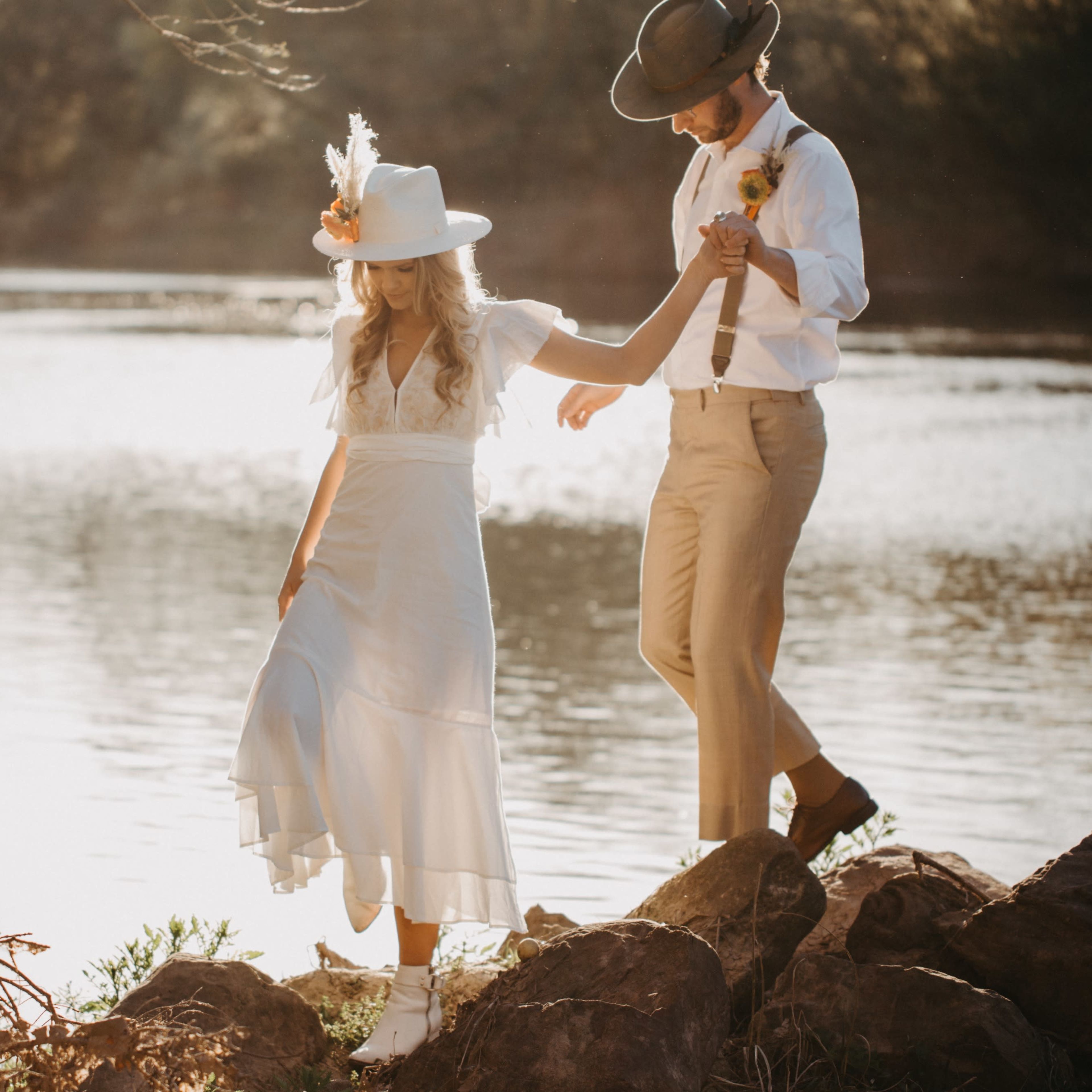 A couple walks hand in hand along the rocky shore of a river, with the sun setting in the background.