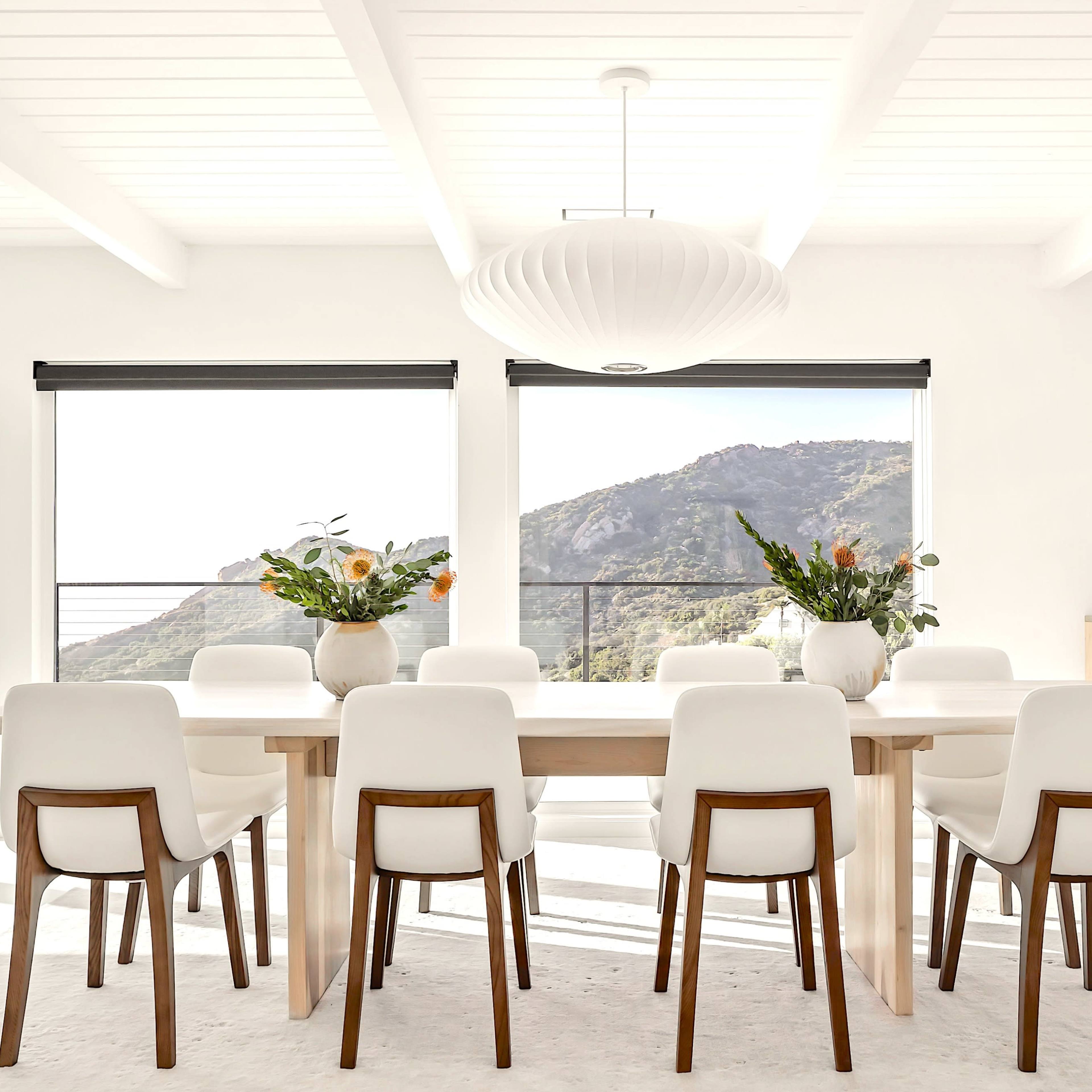 A spacious dining room features a long wooden table surrounded by white chairs, large windows revealing a mountain view, and modern decor.