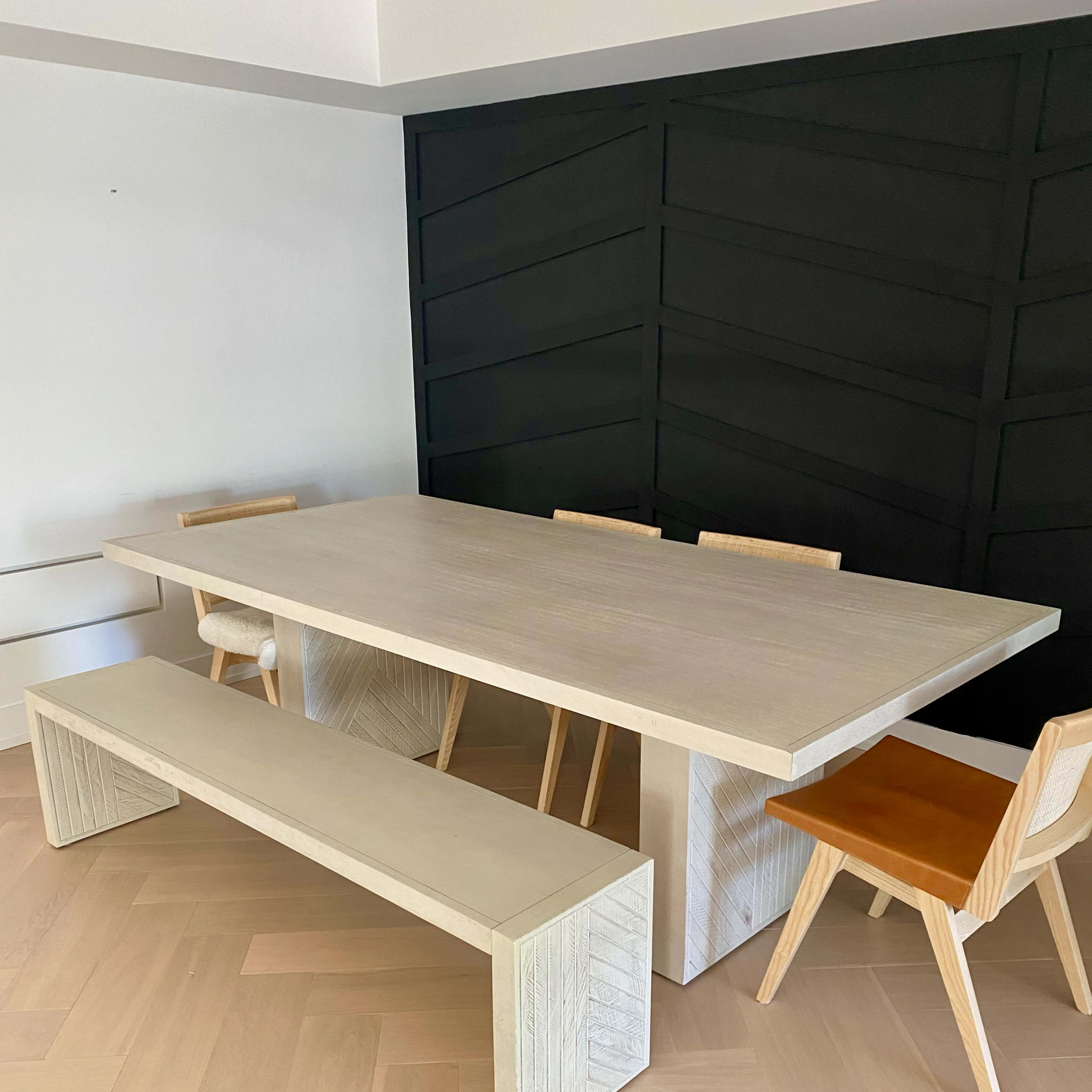 A rectangular dining table with a light wooden finish is surrounded by wooden chairs and a long bench in a modern interior with a dark accent wall.