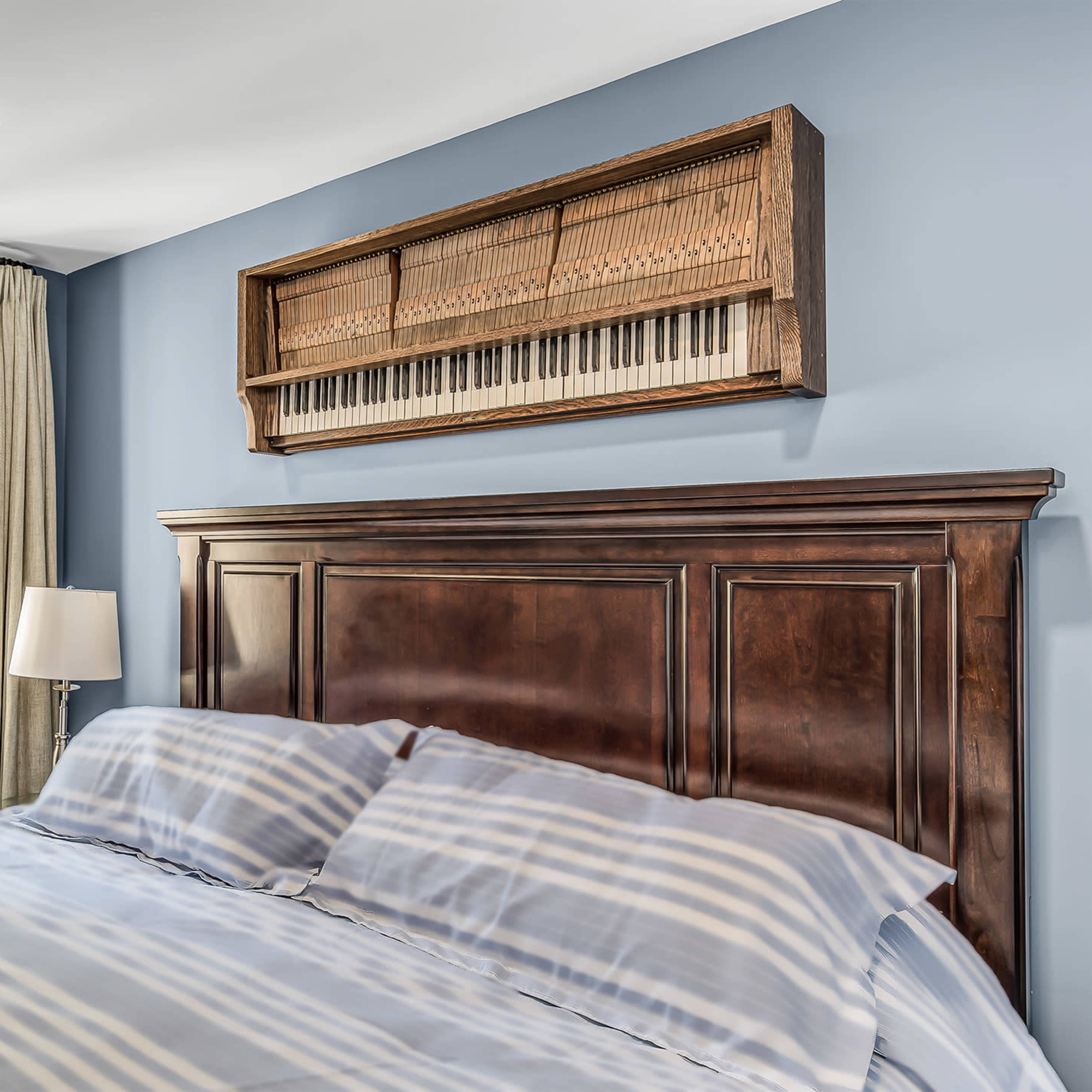 A bedroom with a wooden bed frame, striped bedding, a window with curtains, and a piano keyboard mounted on the wall above the bed.