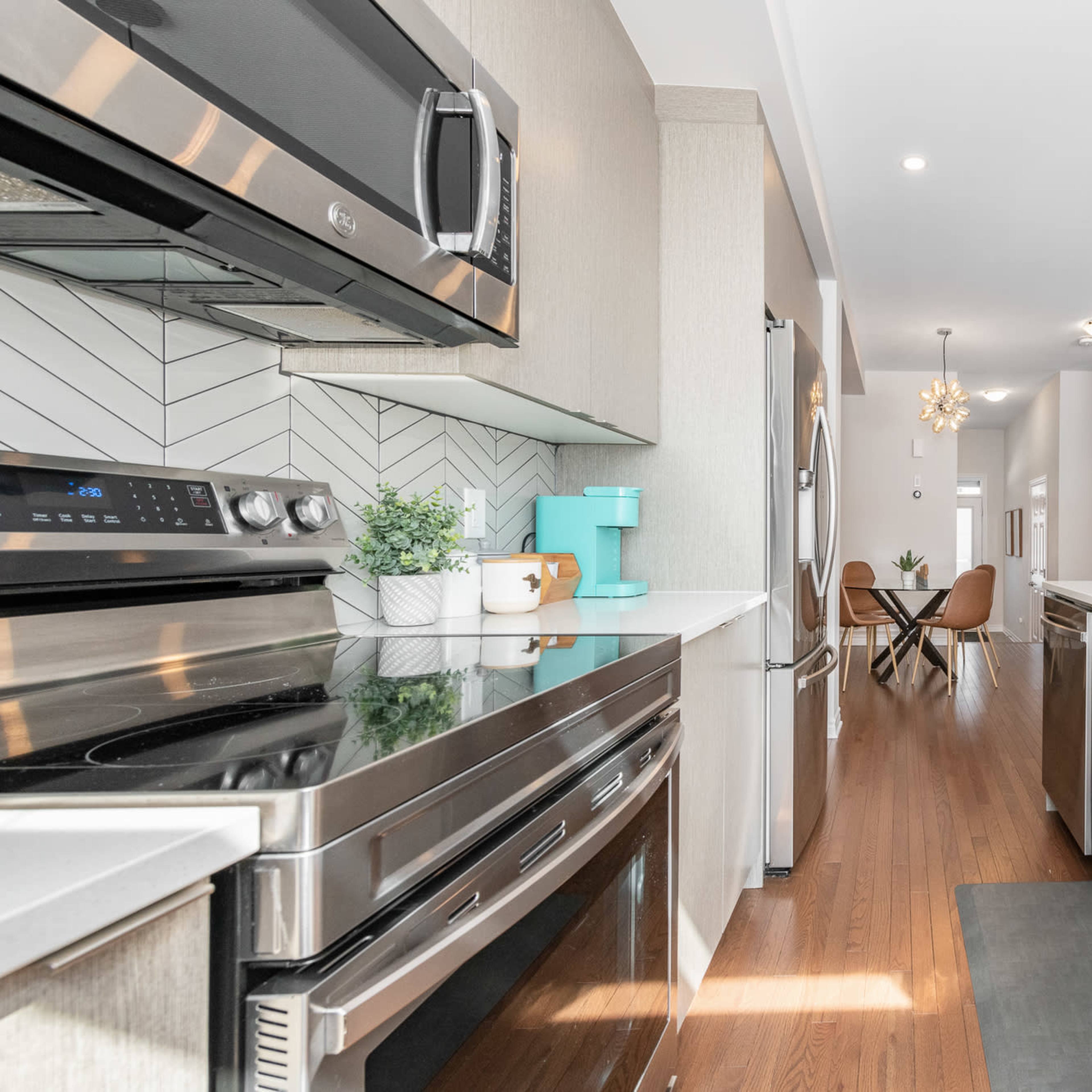 A modern kitchen features stainless steel appliances, a central island with a sink, and light wood cabinetry, leading into a bright dining area.