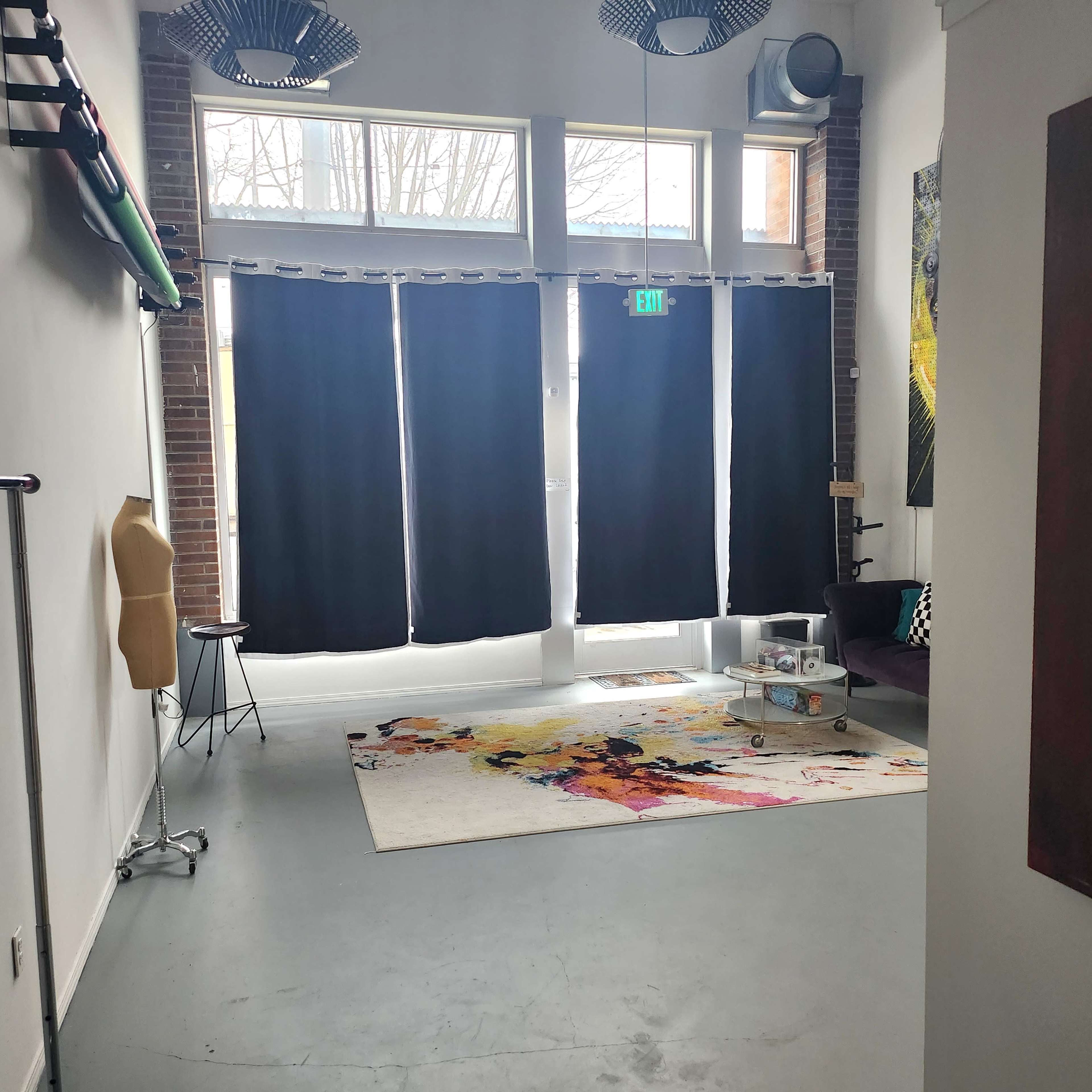 A spacious room with large windows covered by black curtains, a patterned rug on the floor, a clothing rack on the left, and a dark sofa facing a small coffee table.