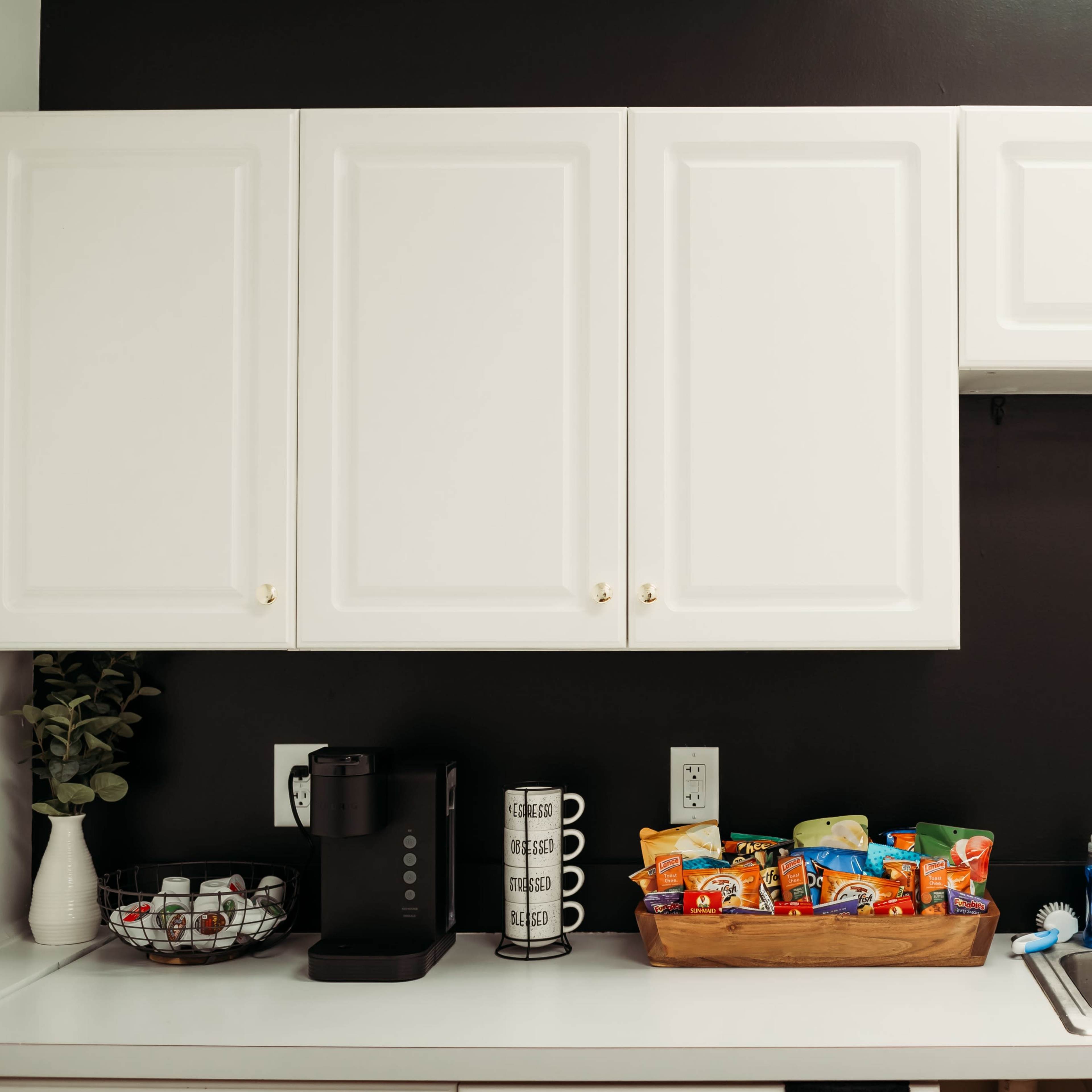 A modern kitchen features white cabinets above a counter with a coffee machine, a bowl of coffee pods, and a basket of snacks beside a sink.