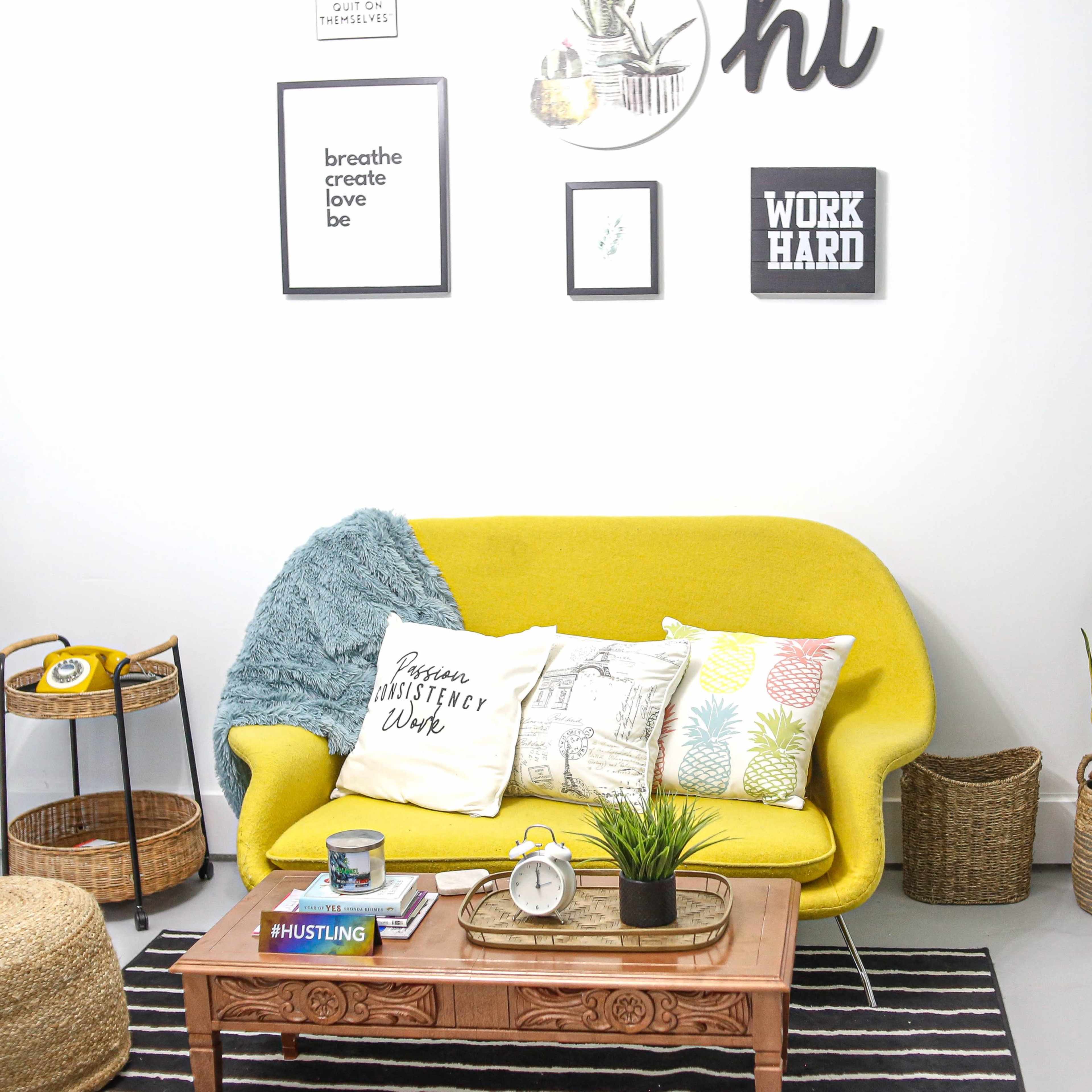 A bright yellow sofa with decorative pillows is positioned in a styled living room featuring a coffee table, plants, and wall art.