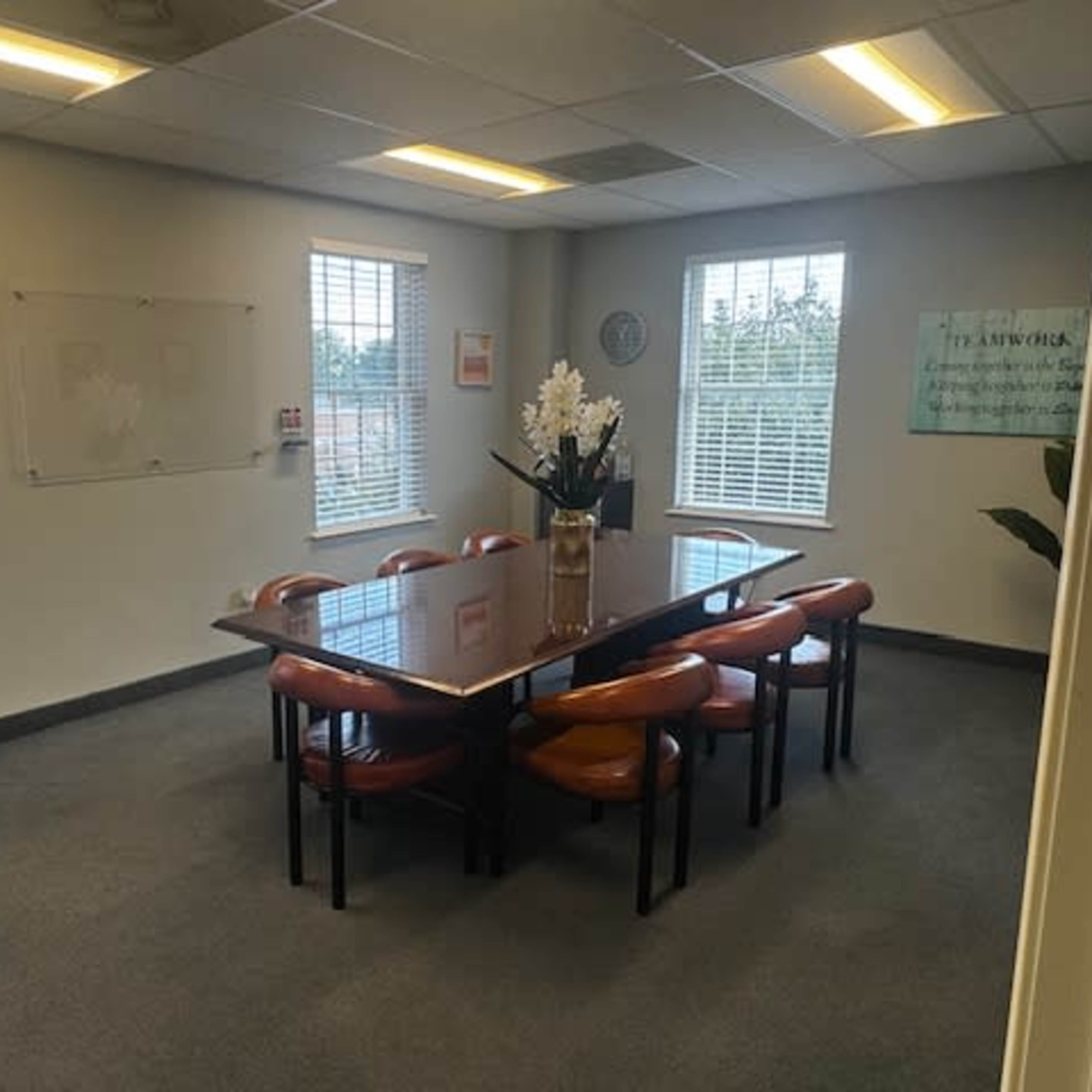 A meeting room features a large table surrounded by several chairs, with windows allowing natural light and a whiteboard on the wall.