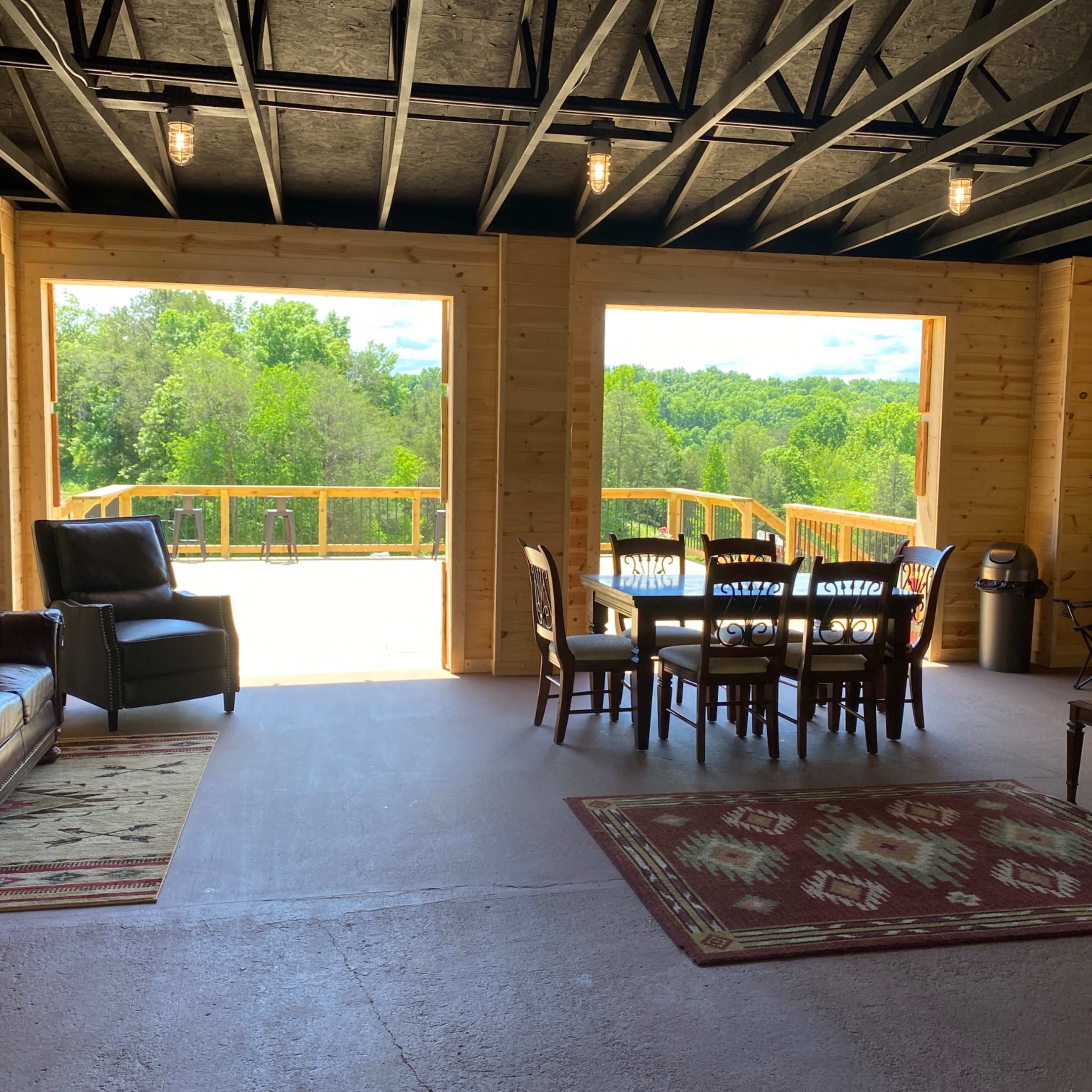 An interior space featuring a leather sofa, wooden dining table with chairs, and large open windows overlooking a green landscape.