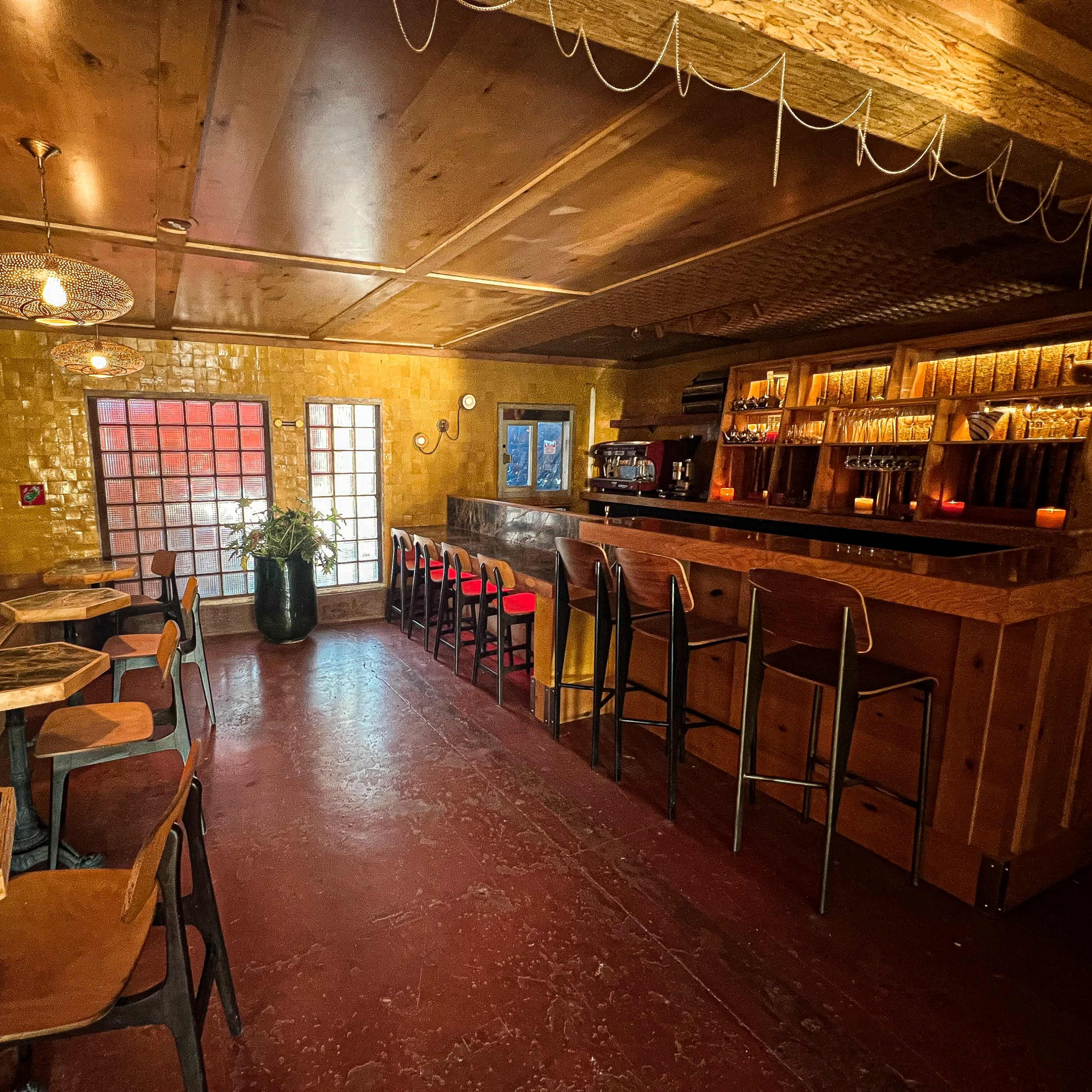 A bar interior with a wooden counter, red accent chairs, and tables, illuminated by warm lighting against a yellow wall.