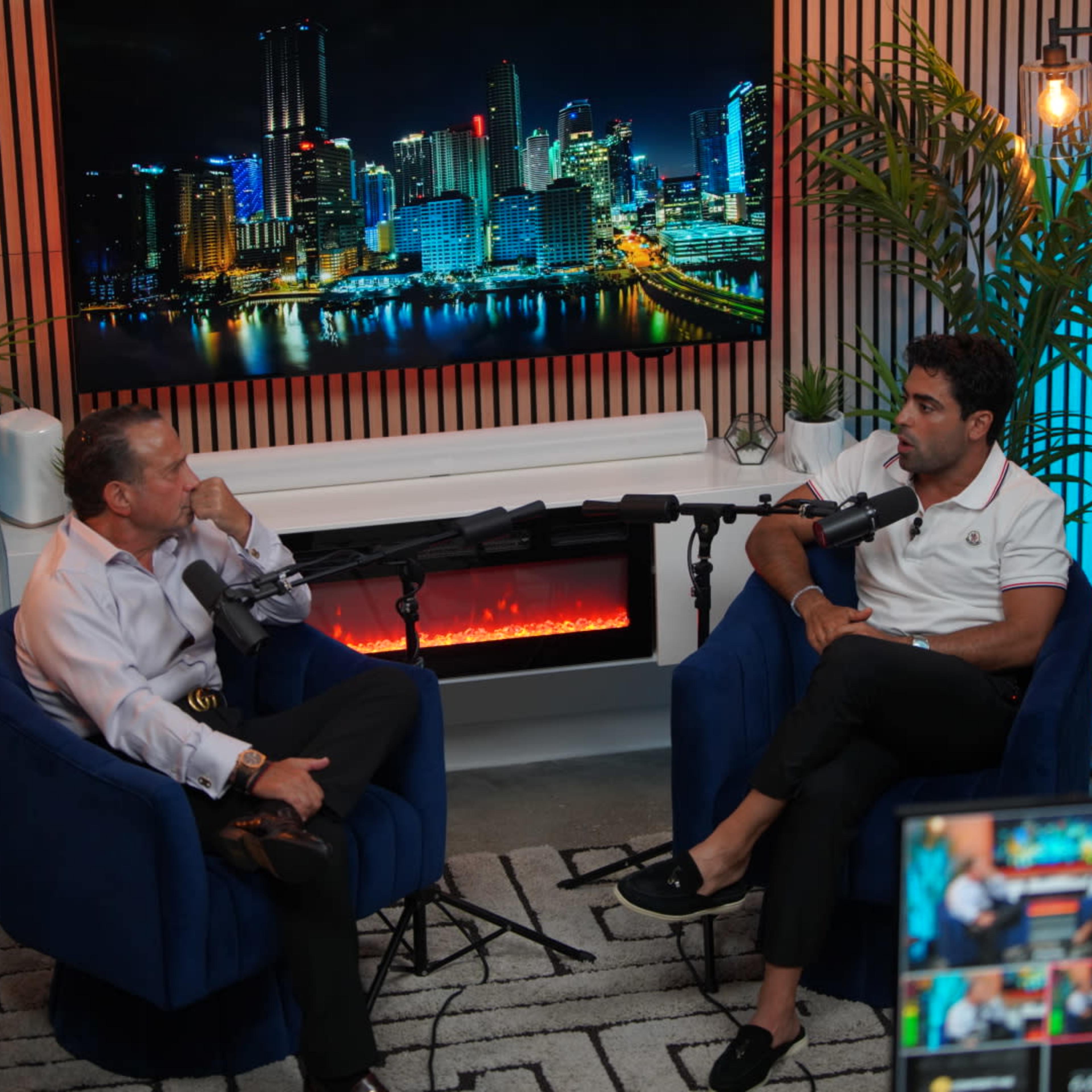 Two men sit in blue chairs in a modern studio, engaged in conversation in front of a large city skyline backdrop.