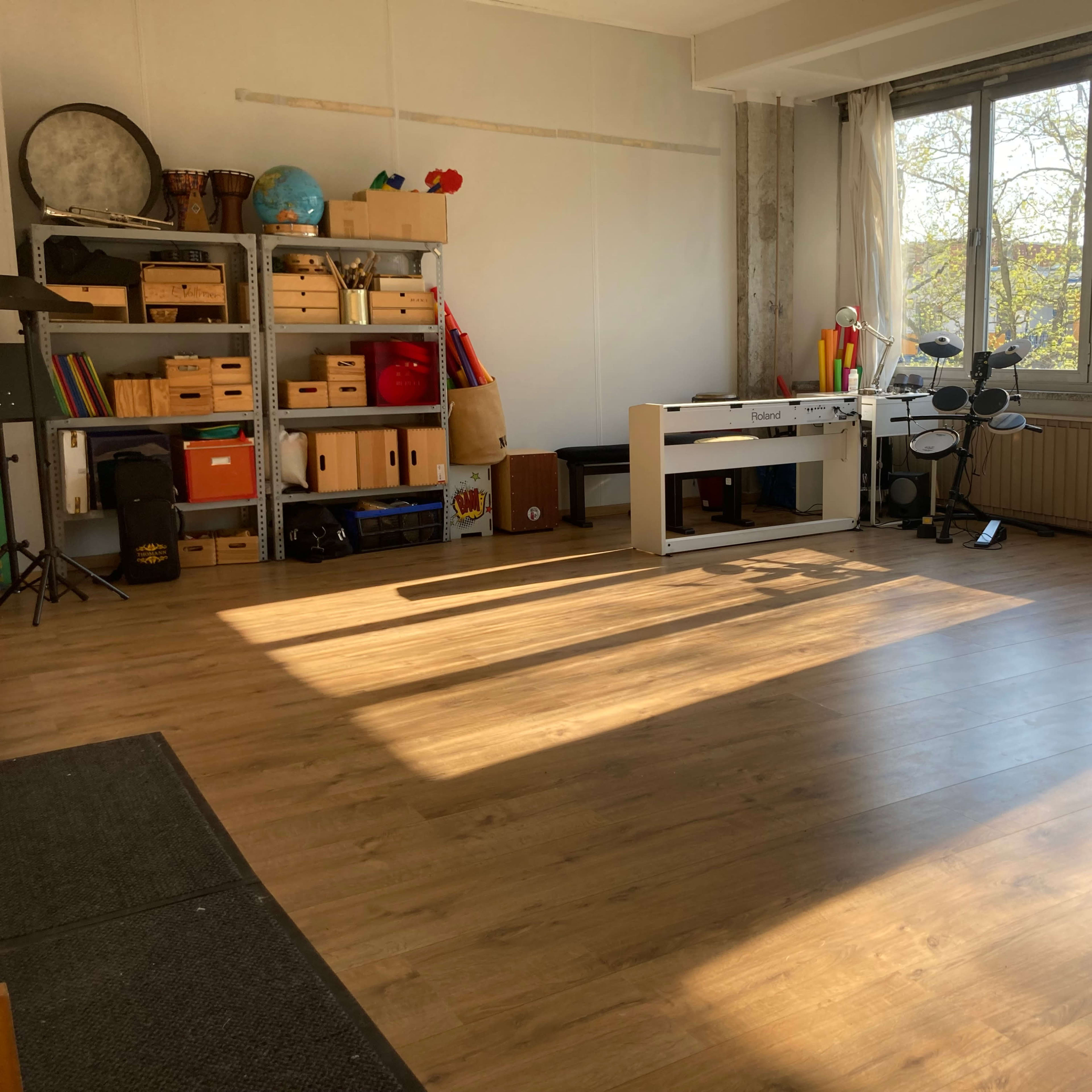 A spacious room with wooden floors, bright sunlight streaming in through large windows, and various storage boxes and musical instruments against the walls.