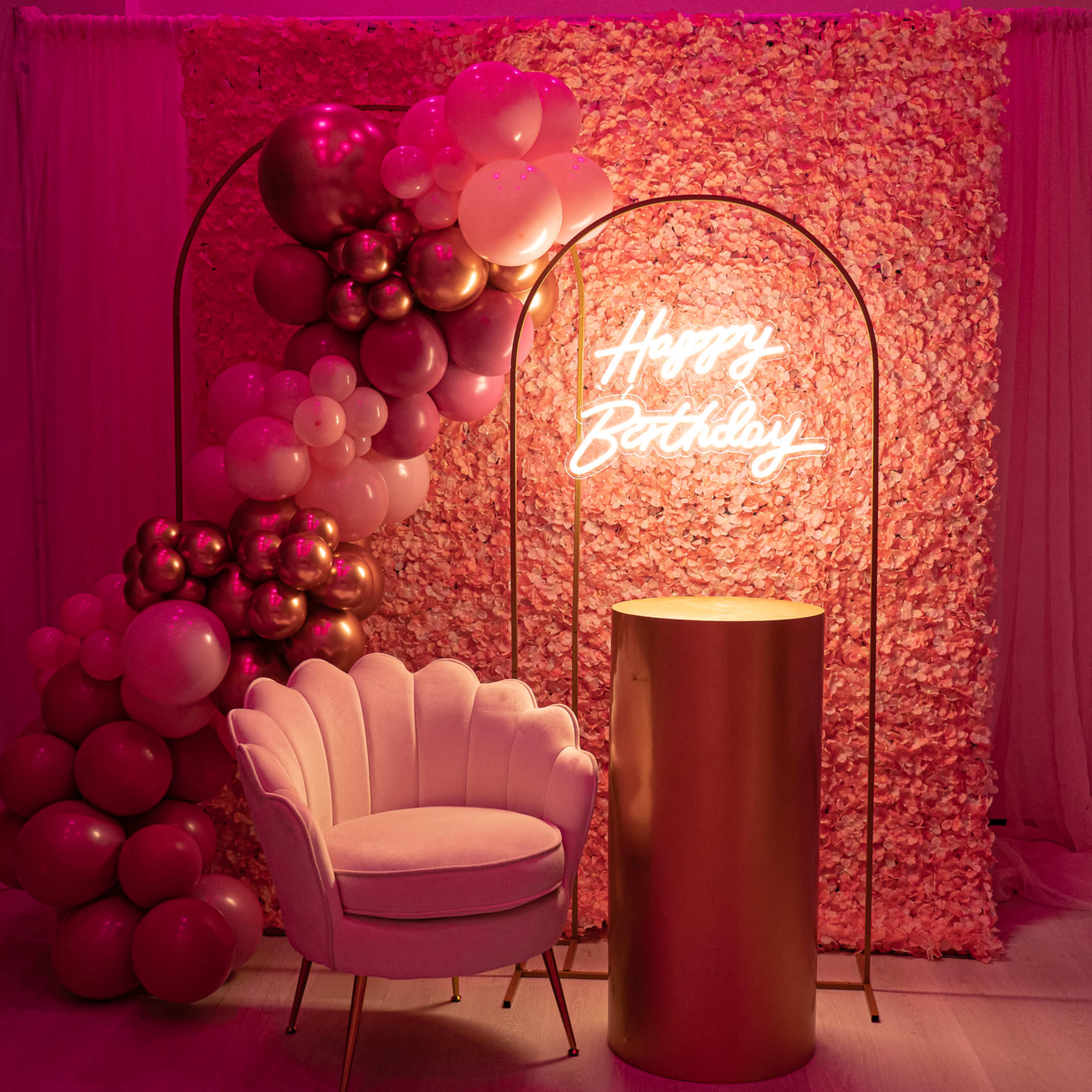 A festive birthday decoration featuring a pink balloon arrangement, a floral backdrop, and a glowing "Happy Birthday" sign next to a white chair and a gold table.