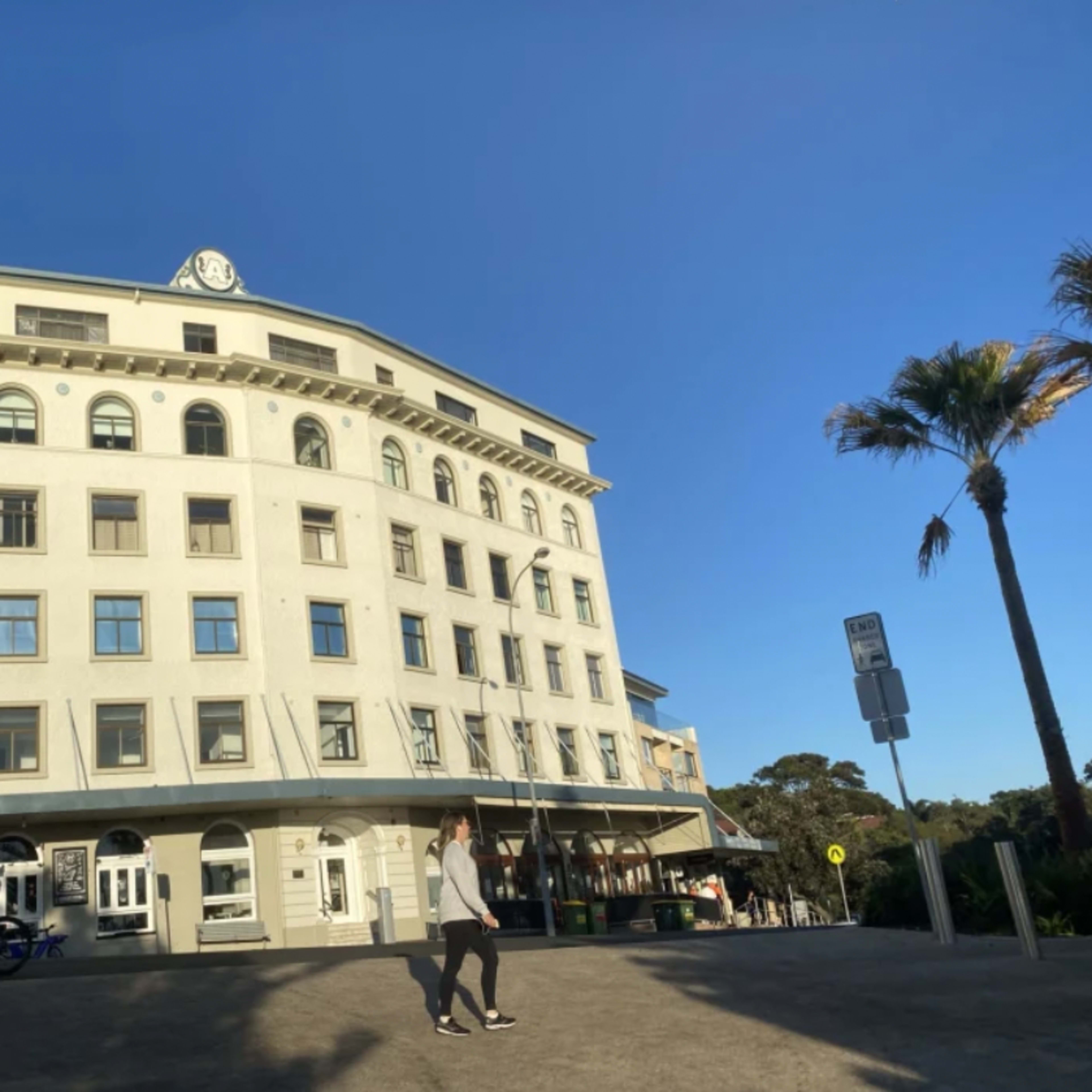 A person walks past a historic building with a clock on the roof, located in a coastal area with palm trees.