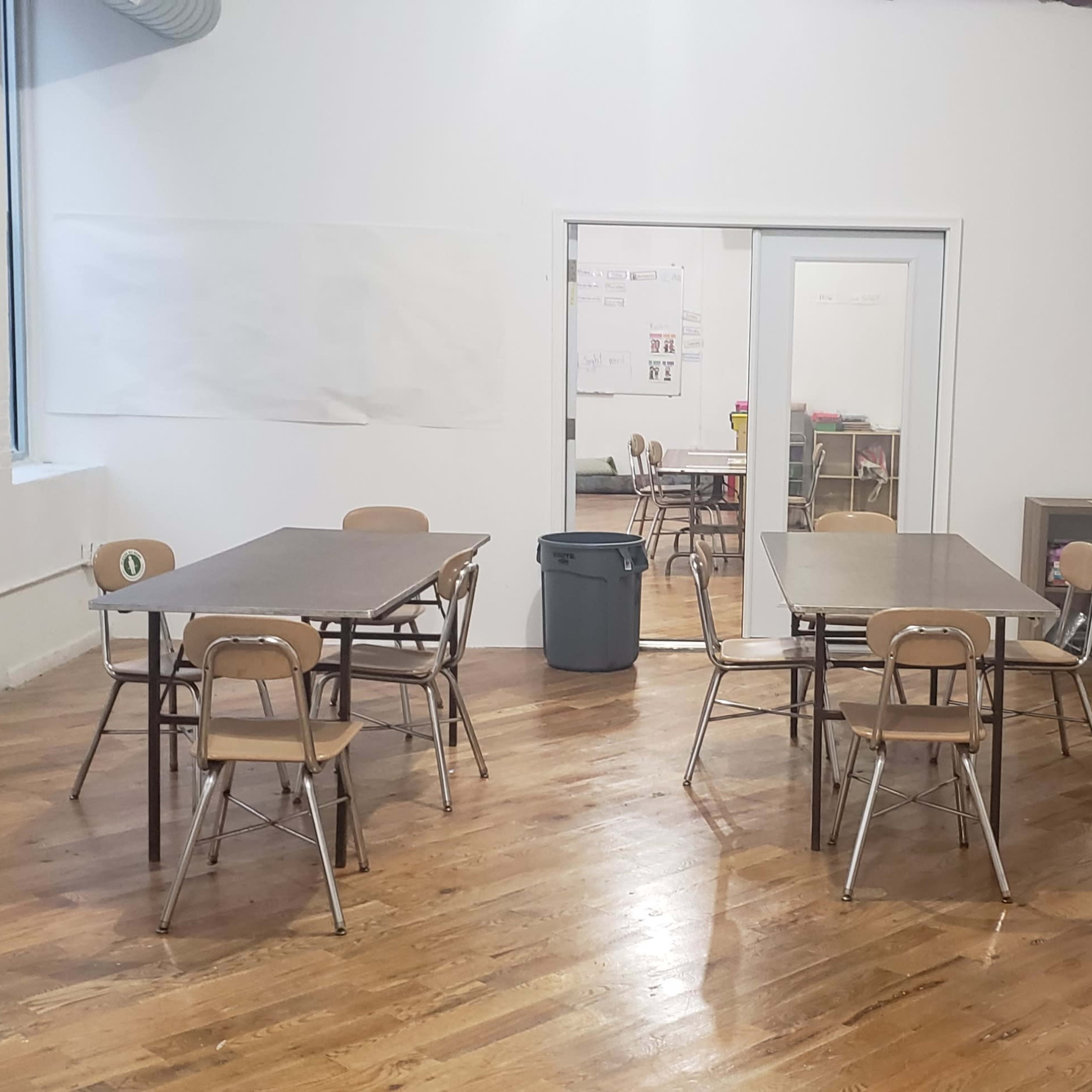 An empty classroom with several wooden tables and chairs arranged around a hardwood floor, along with a storage shelf and a trash can.