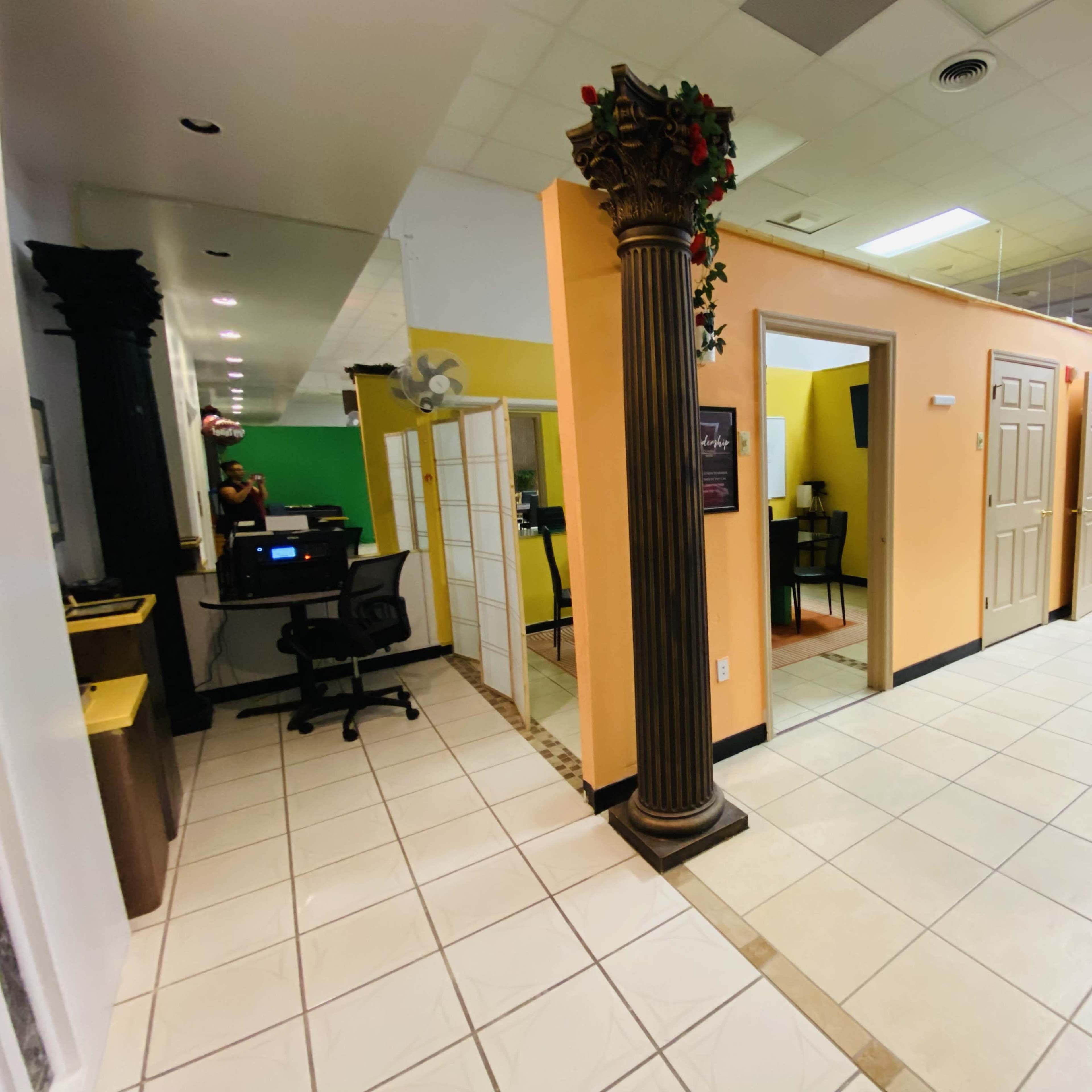 A hallway with several closed doors on the right and an office area with a computer on the left, framed by a decorative column.