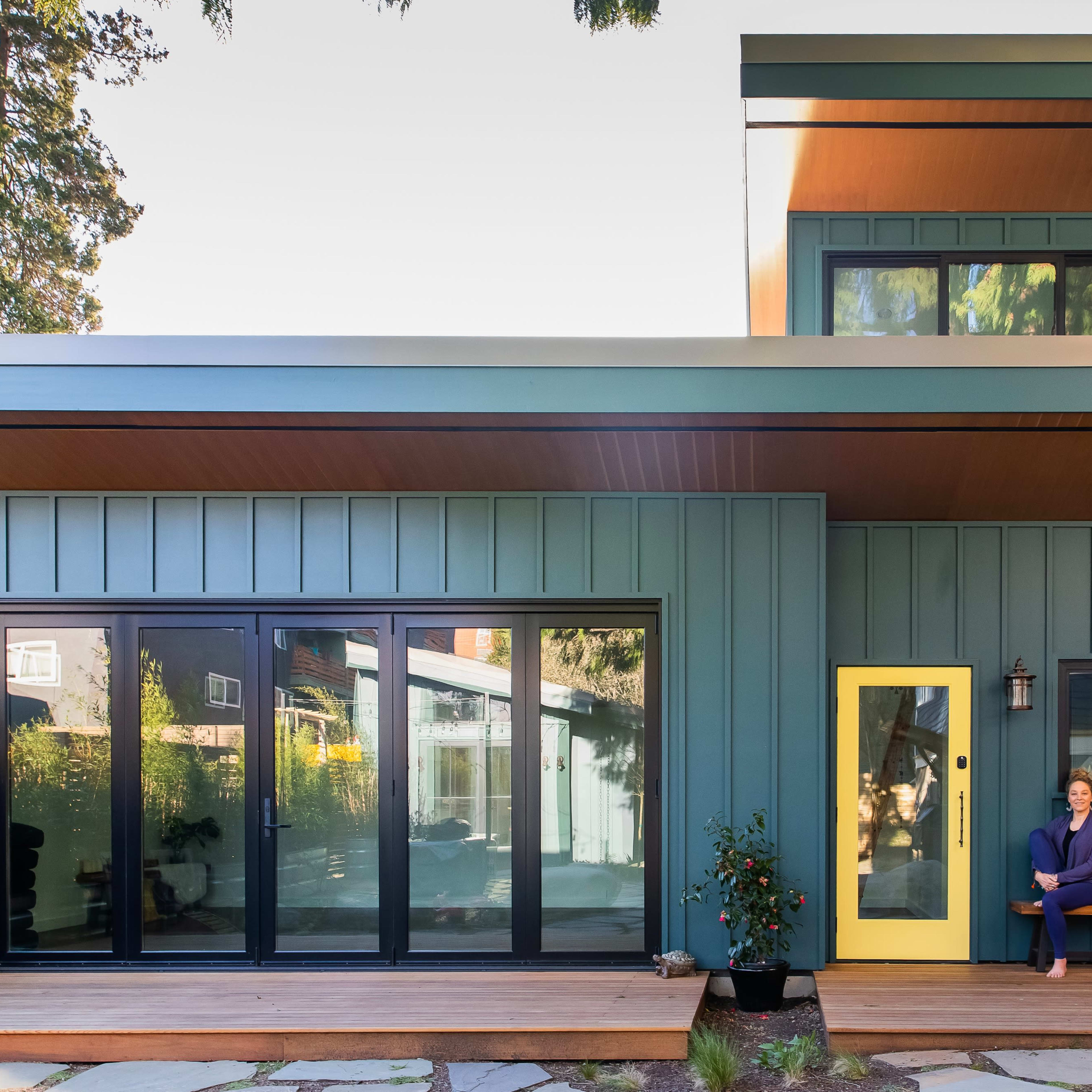 A contemporary house with dark blue siding, large glass doors, and a bright yellow front door features a person sitting on a wooden bench.