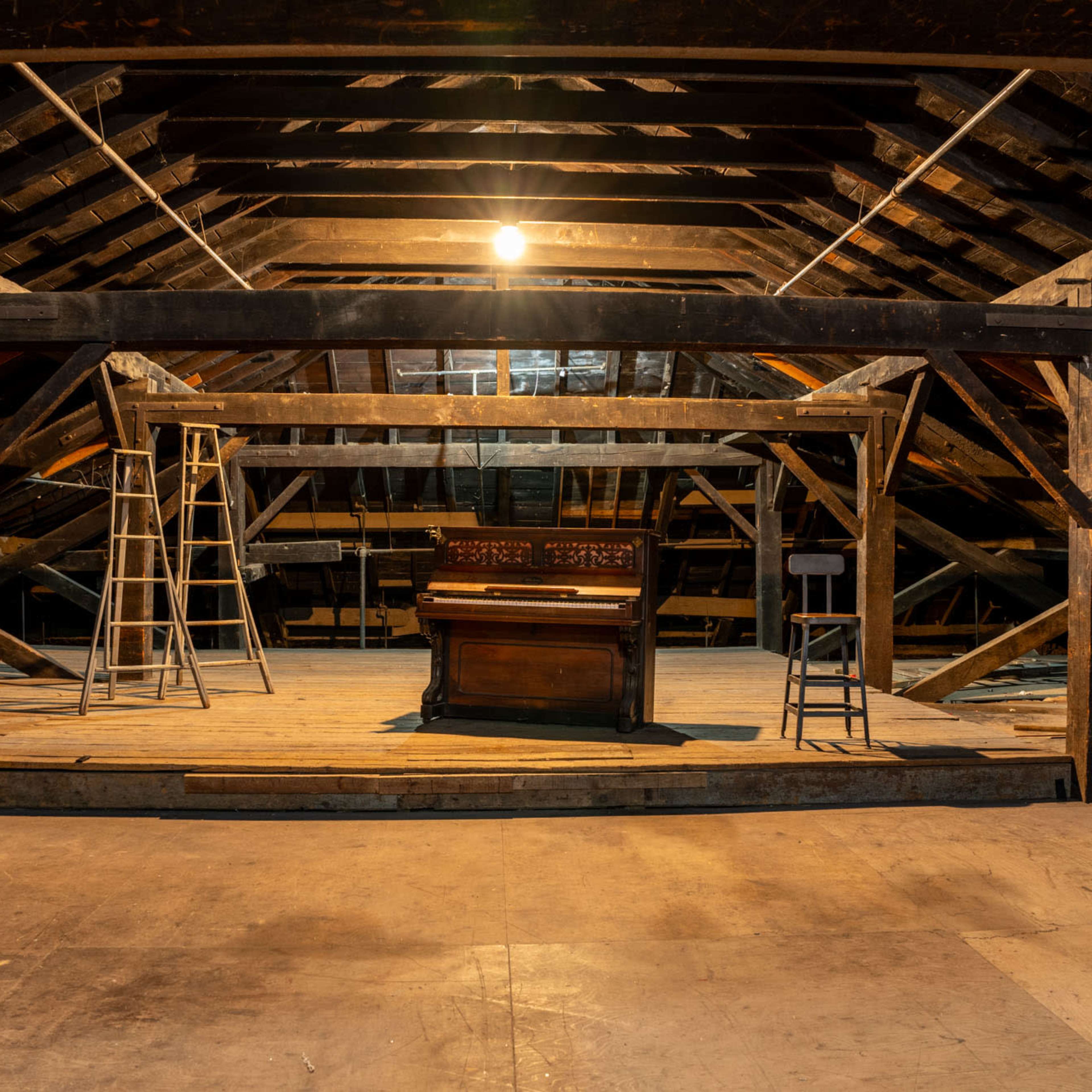 An antique piano is positioned in the center of a spacious, unfinished attic illuminated by a single light.