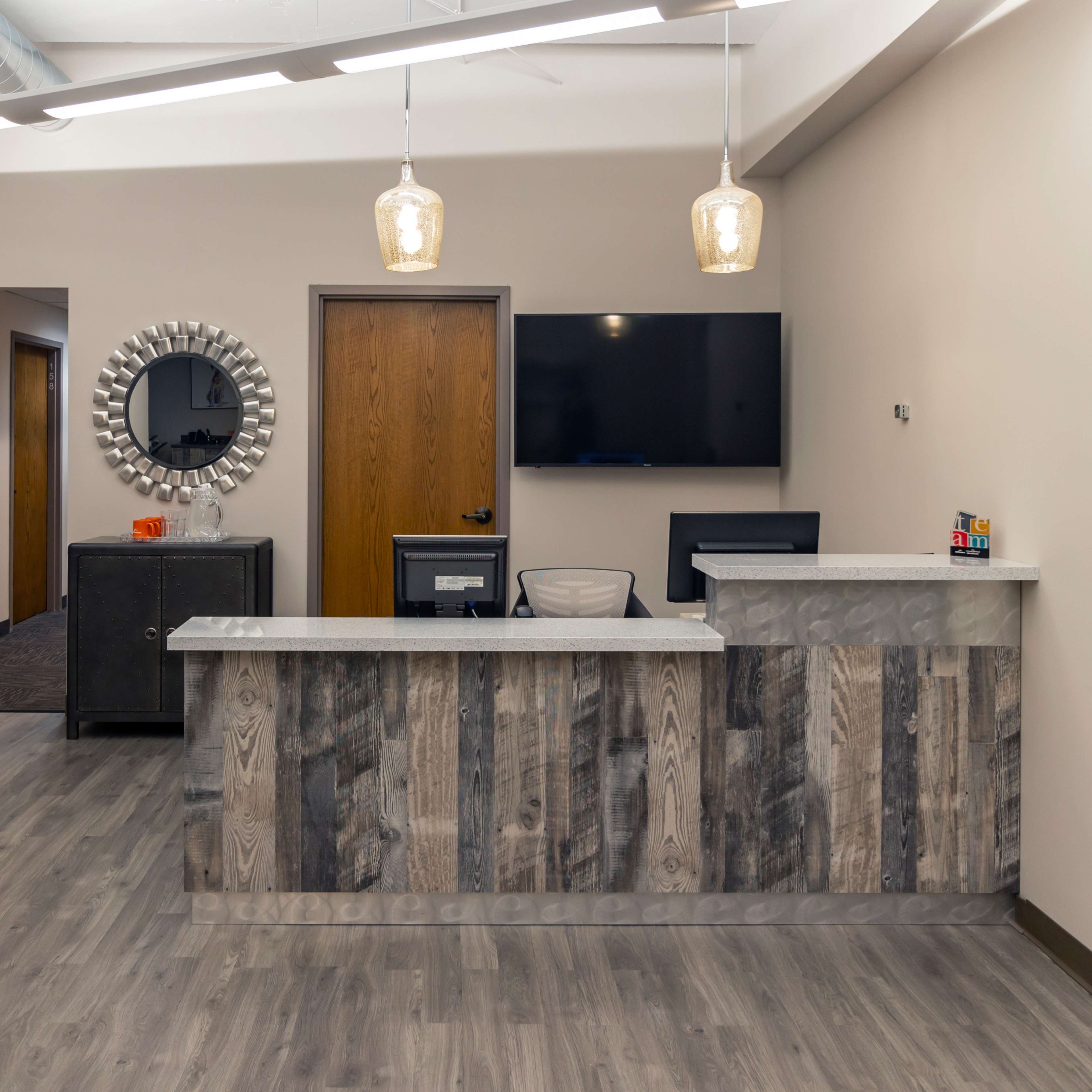 A modern reception area featuring a wooden front desk, a large round mirror, and a wall-mounted TV in a well-lit space.