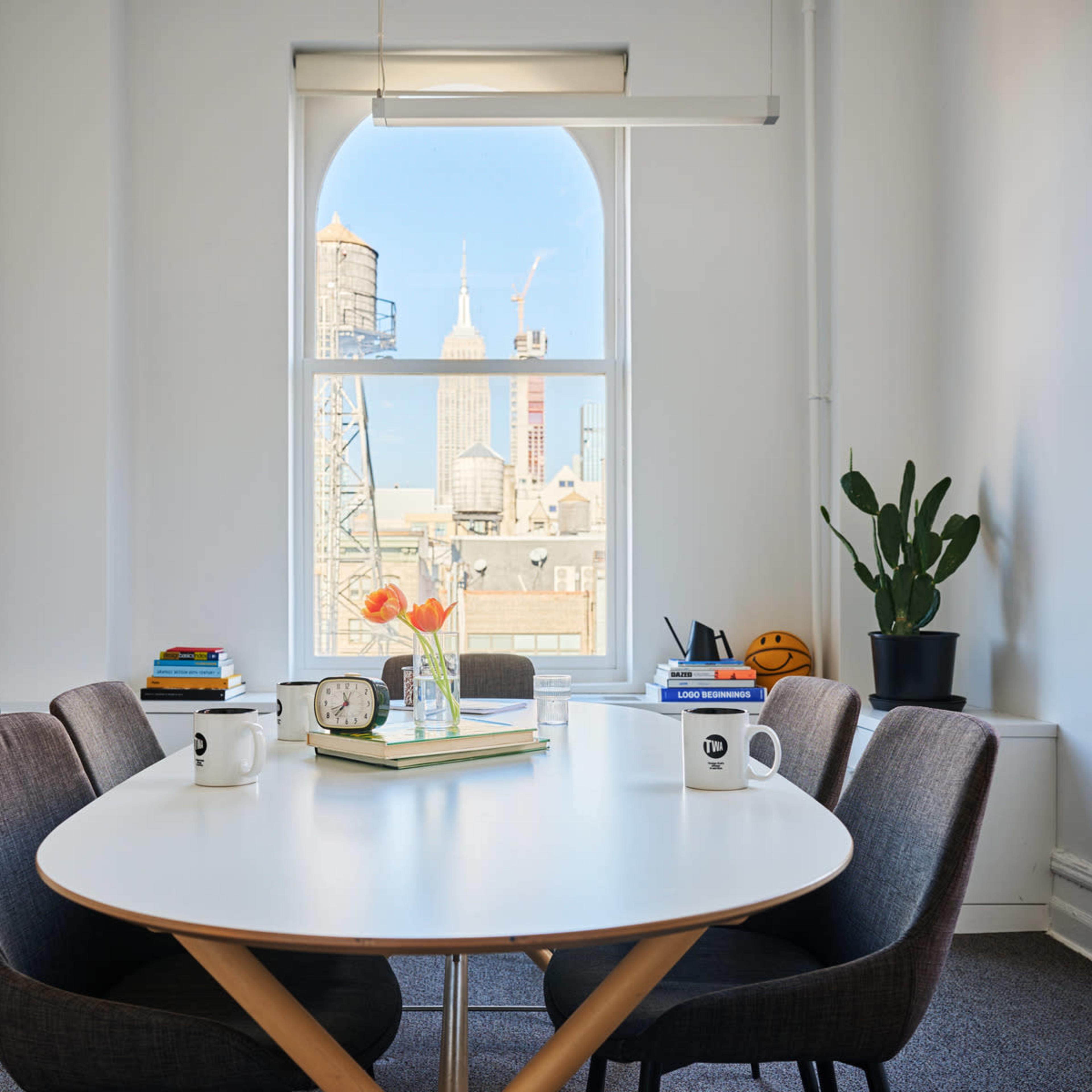 A modern conference room features a circular table surrounded by chairs, with a view of the Empire State Building visible through a large window.