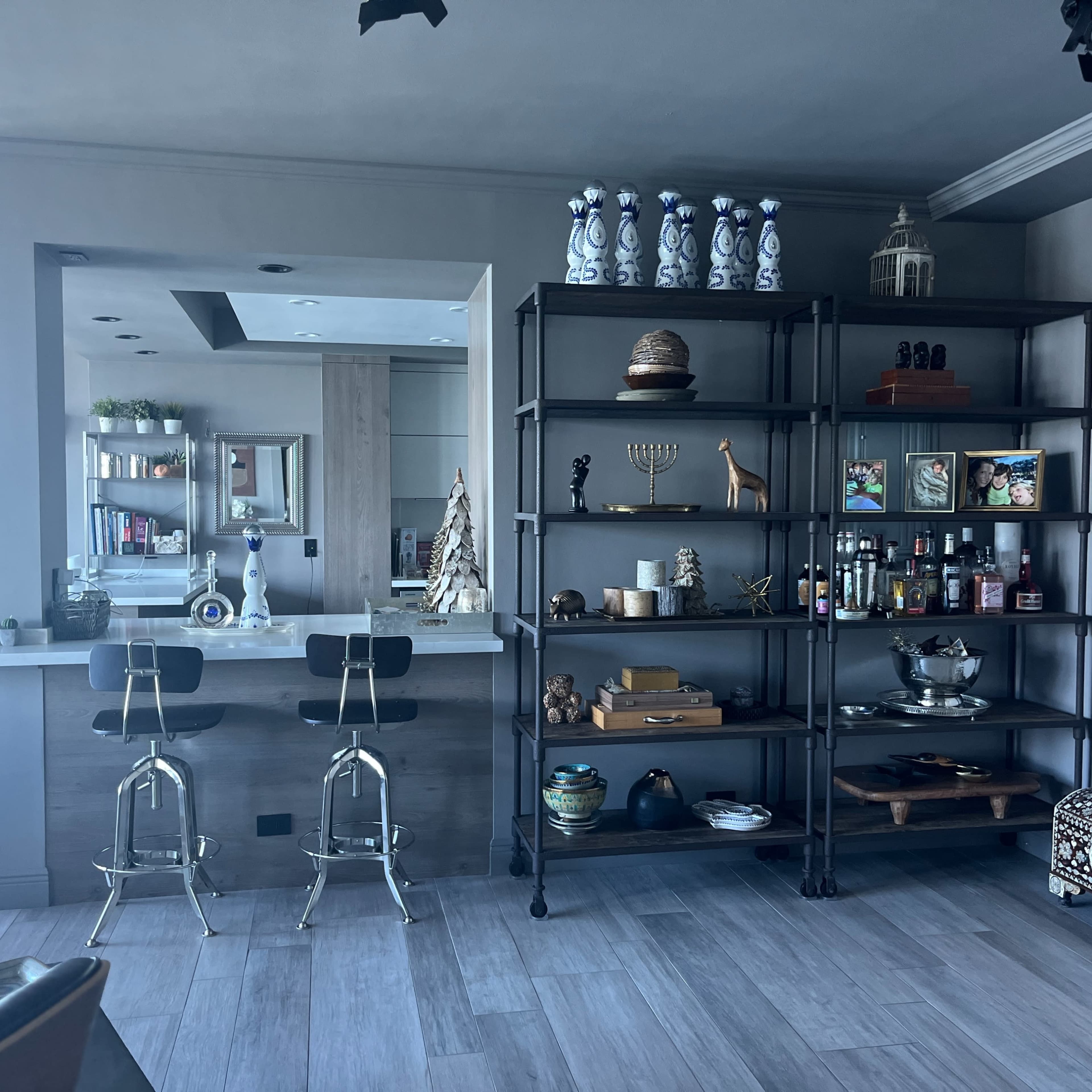 A modern interior features a kitchen bar with two stools and a shelving unit displaying various decorative items and bottles.