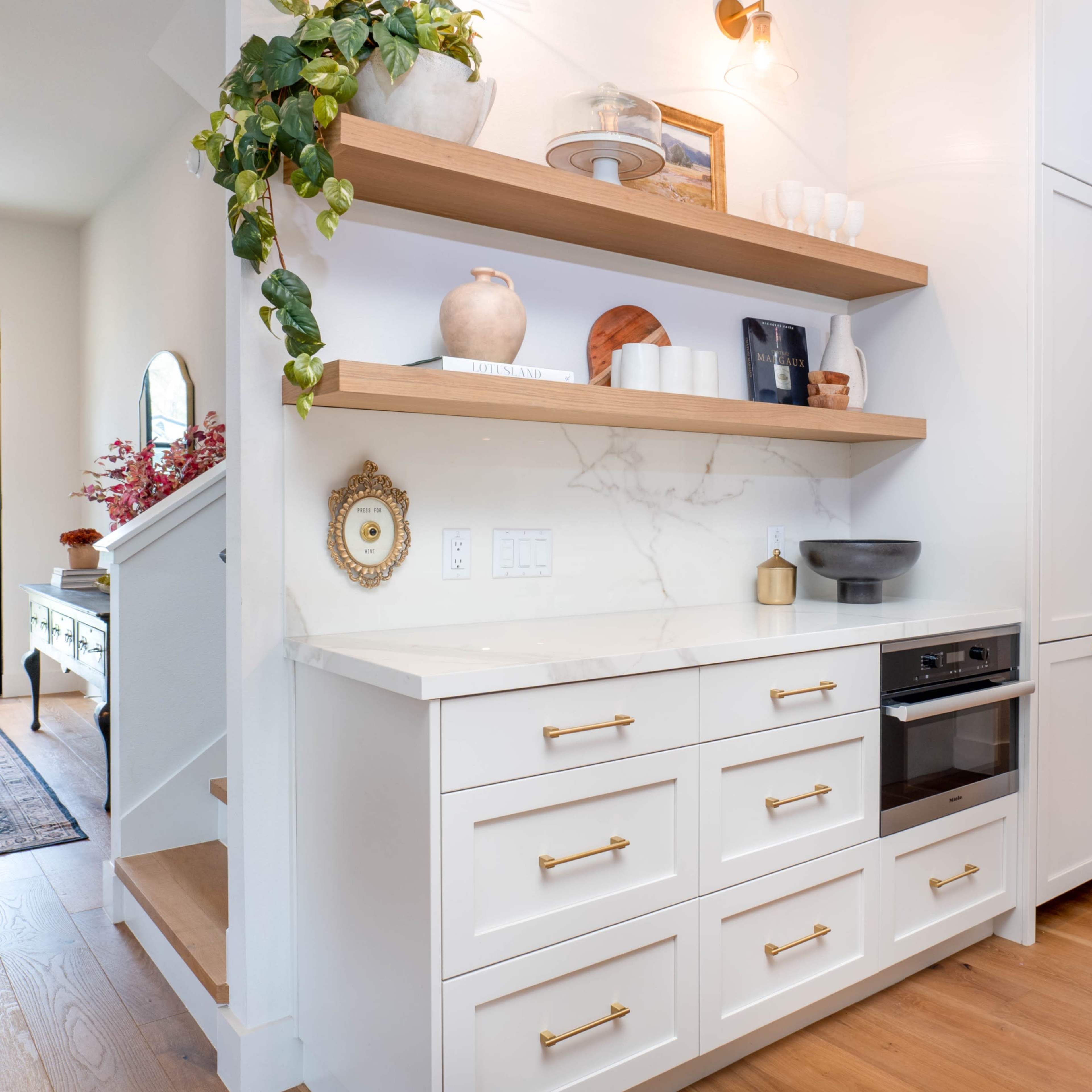 A modern kitchen with white cabinetry, open wooden shelves holding decorative items, and a built-in oven alongside a marble countertop.
