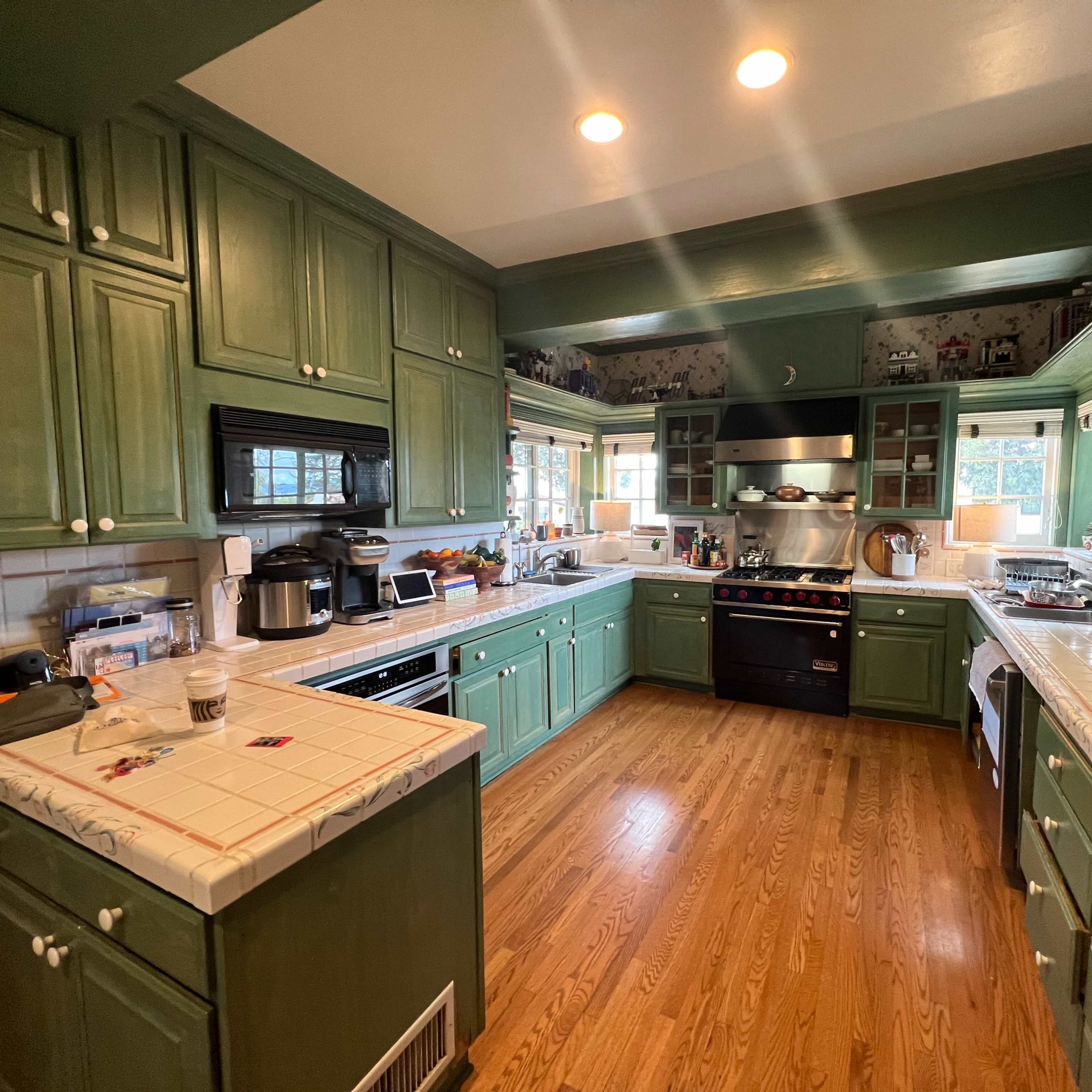 A spacious kitchen with green cabinetry, stainless steel appliances, and a tile countertop, featuring wooden flooring and multiple windows.