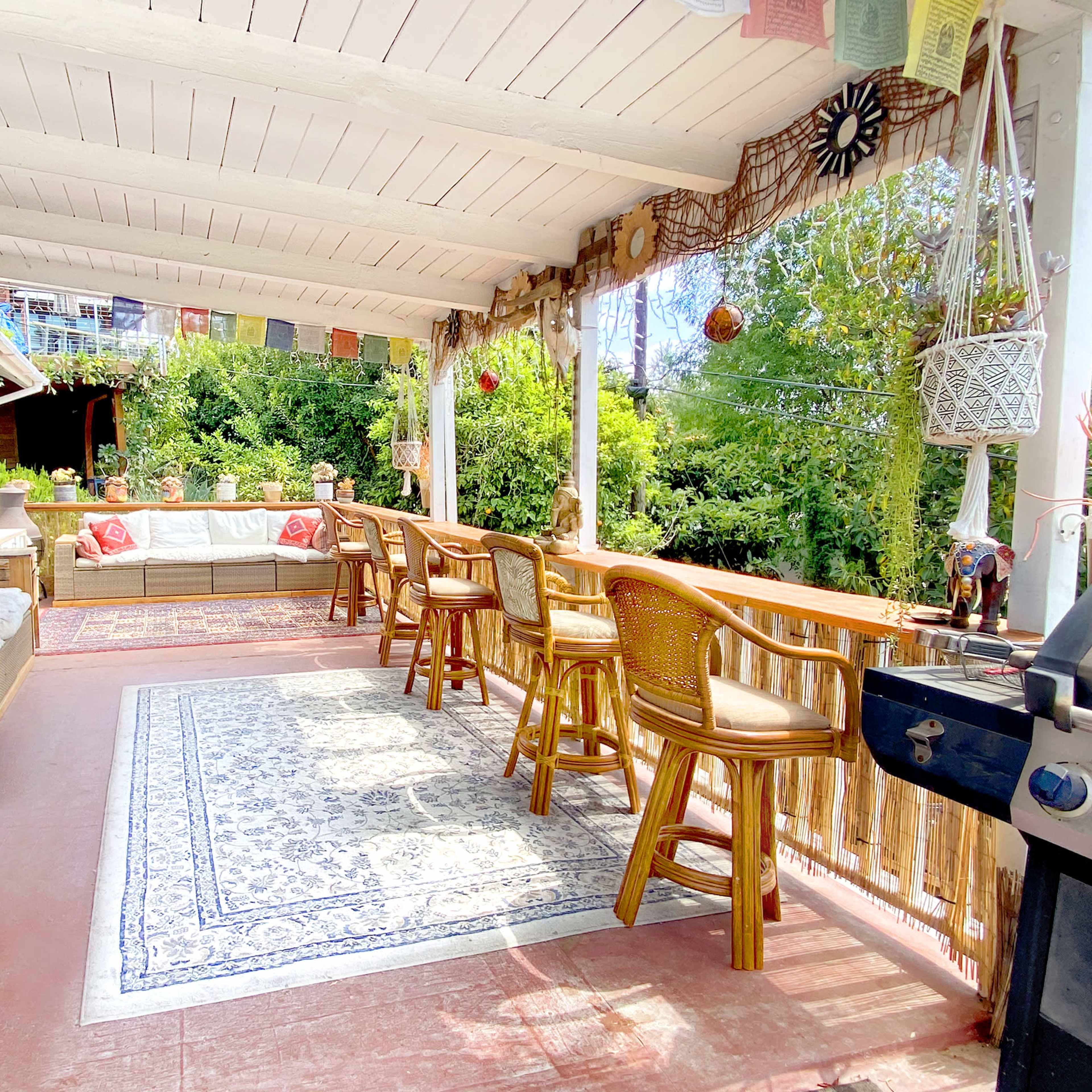 A spacious porch with a seating area, wooden bar stools, a BBQ grill, and greenery visible in the background.
