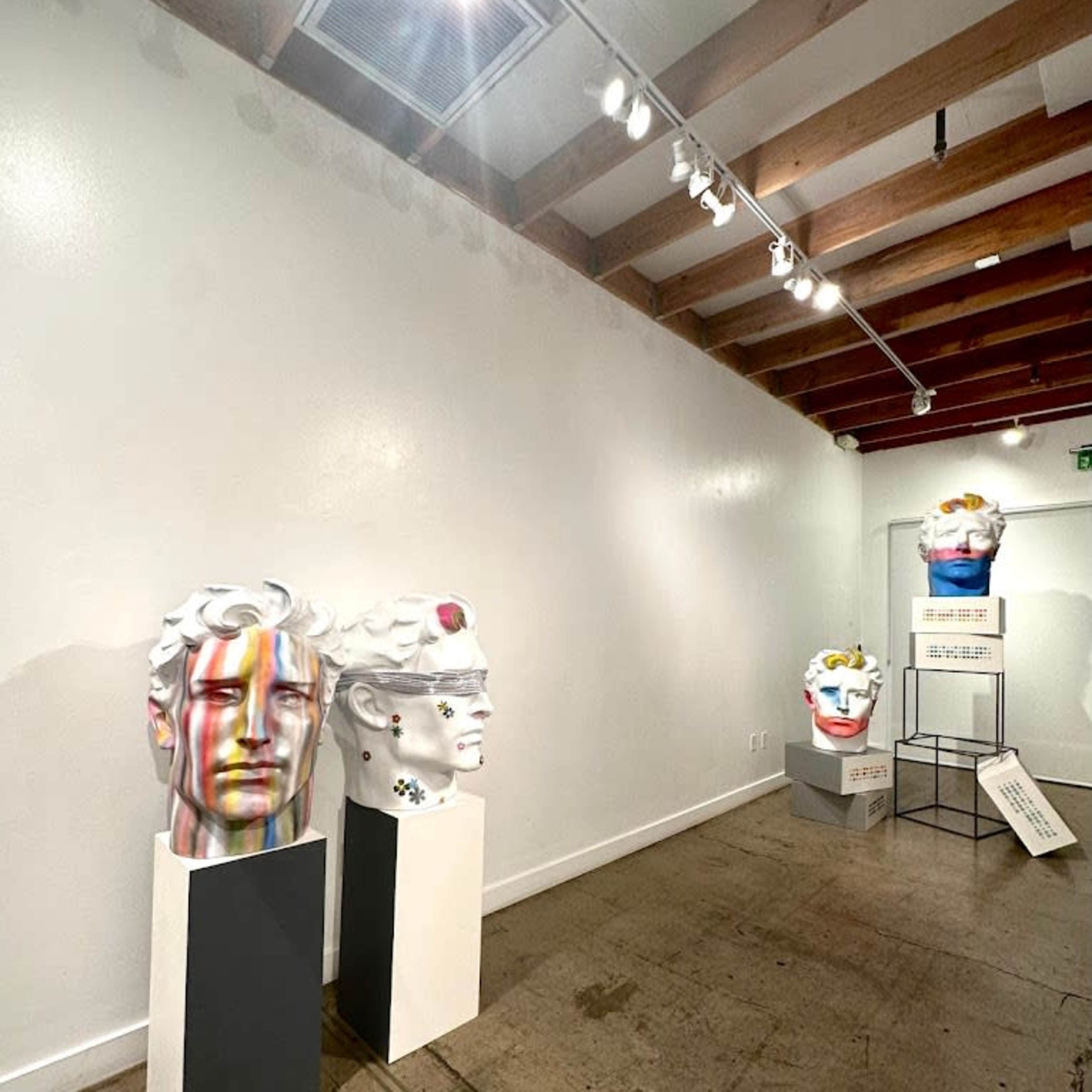 A gallery exhibition featuring several colorful sculptures of human heads displayed on pedestals against a white wall.