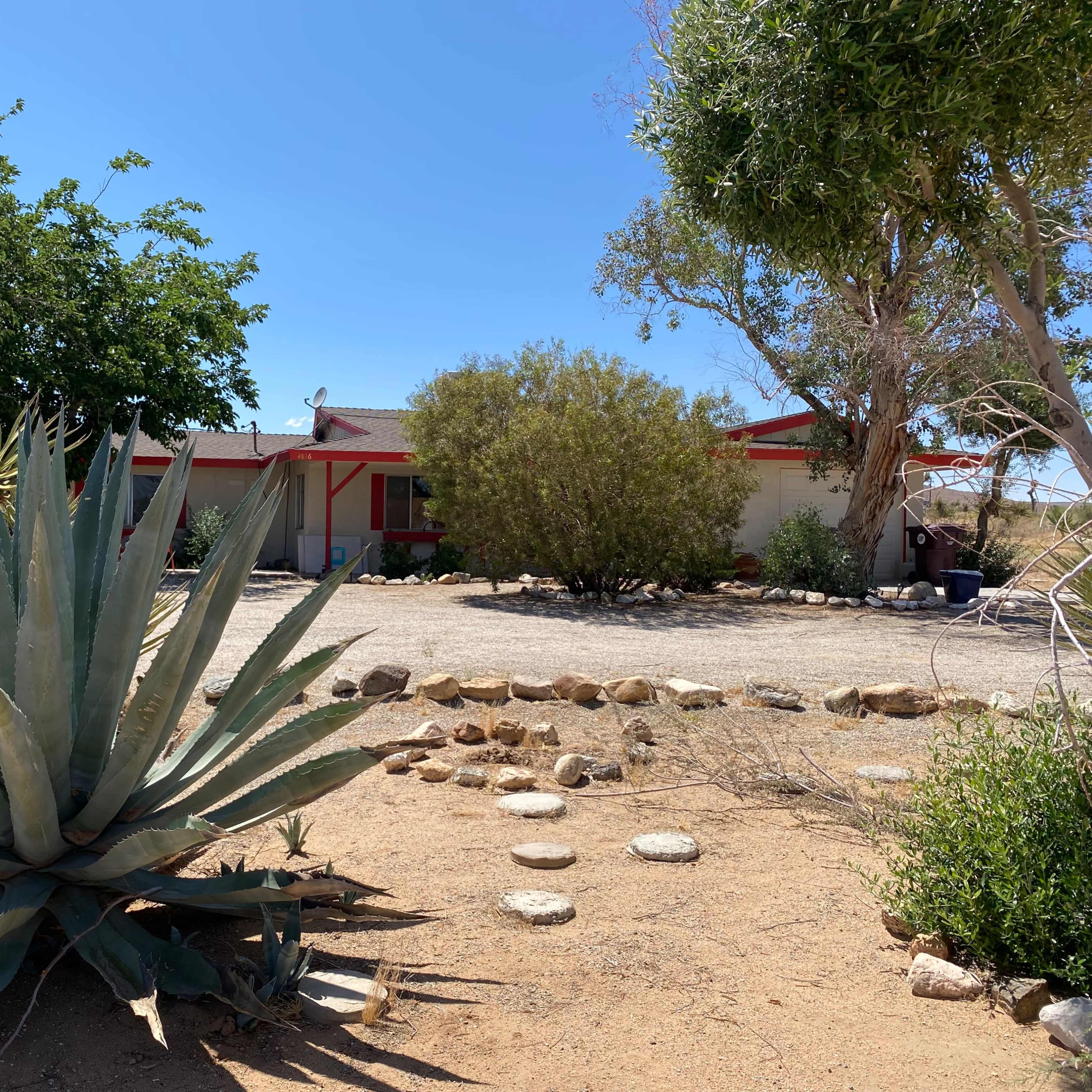 A large agave plant is foregrounded in a desert landscape with a single-story house and sparse vegetation in the background.