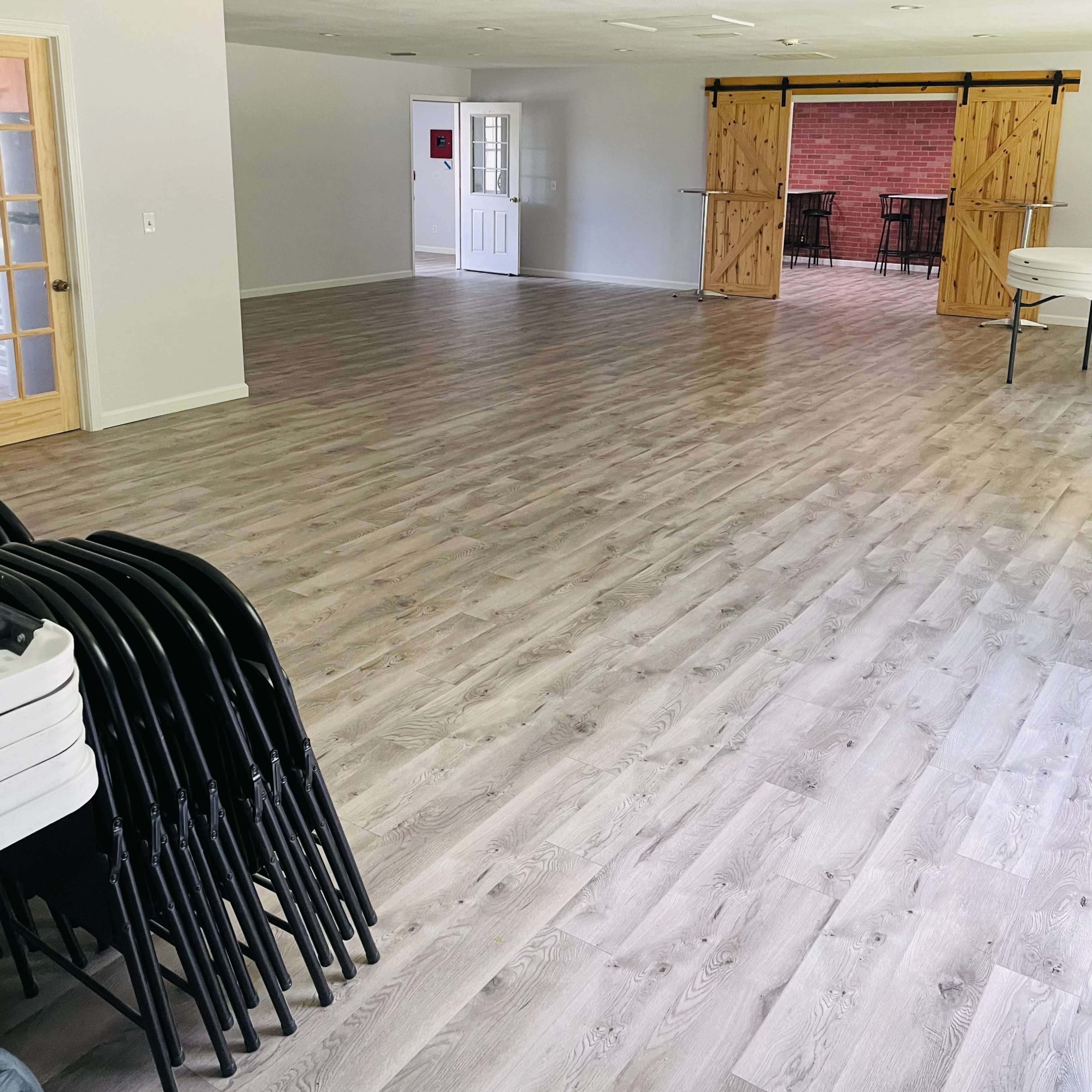 A spacious interior room with light wooden flooring, empty walls, and a set of sliding barn doors leading to a brick patio area.