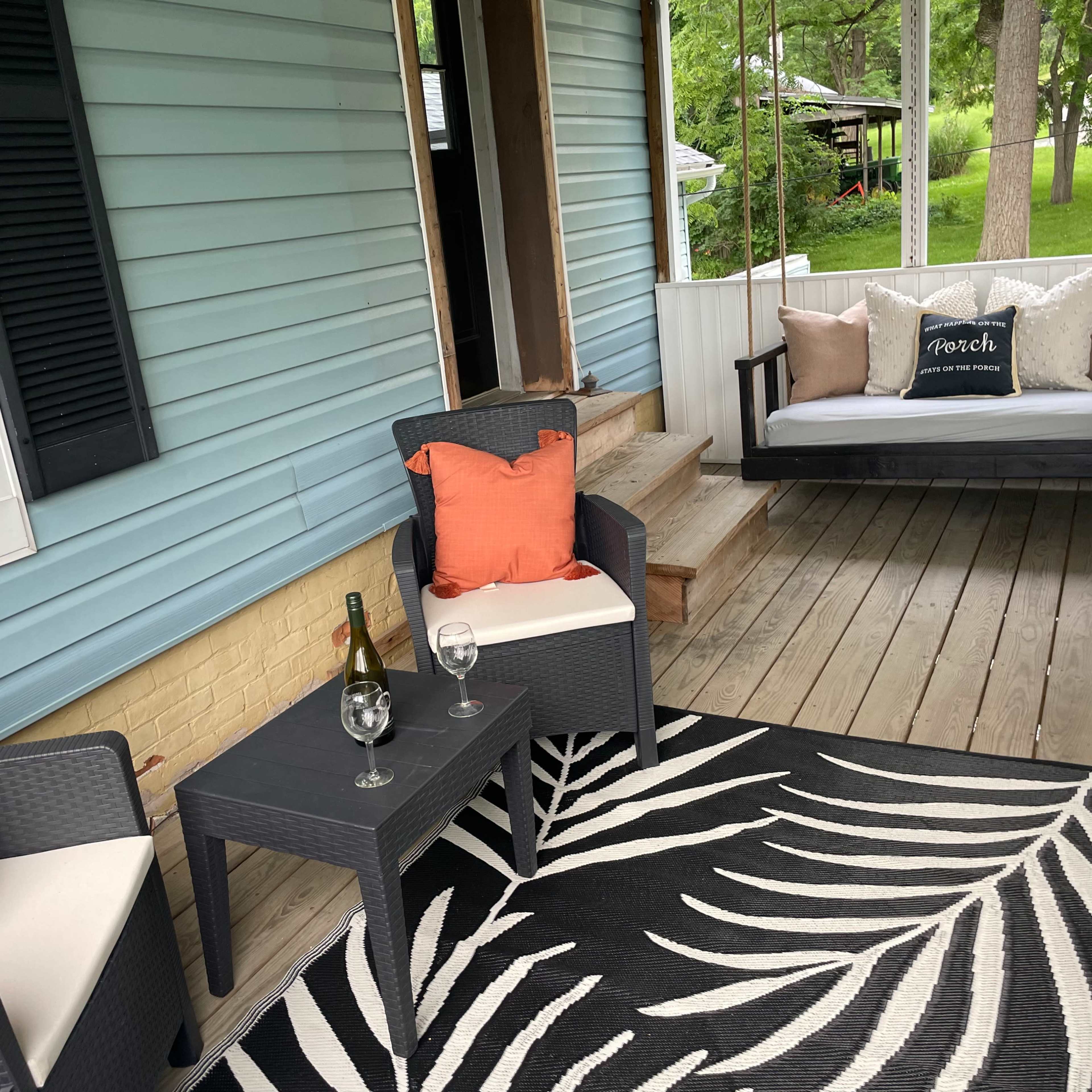 A screened porch with two chairs and a table, a swing with decorative pillows, and a decorative rug on wooden flooring.