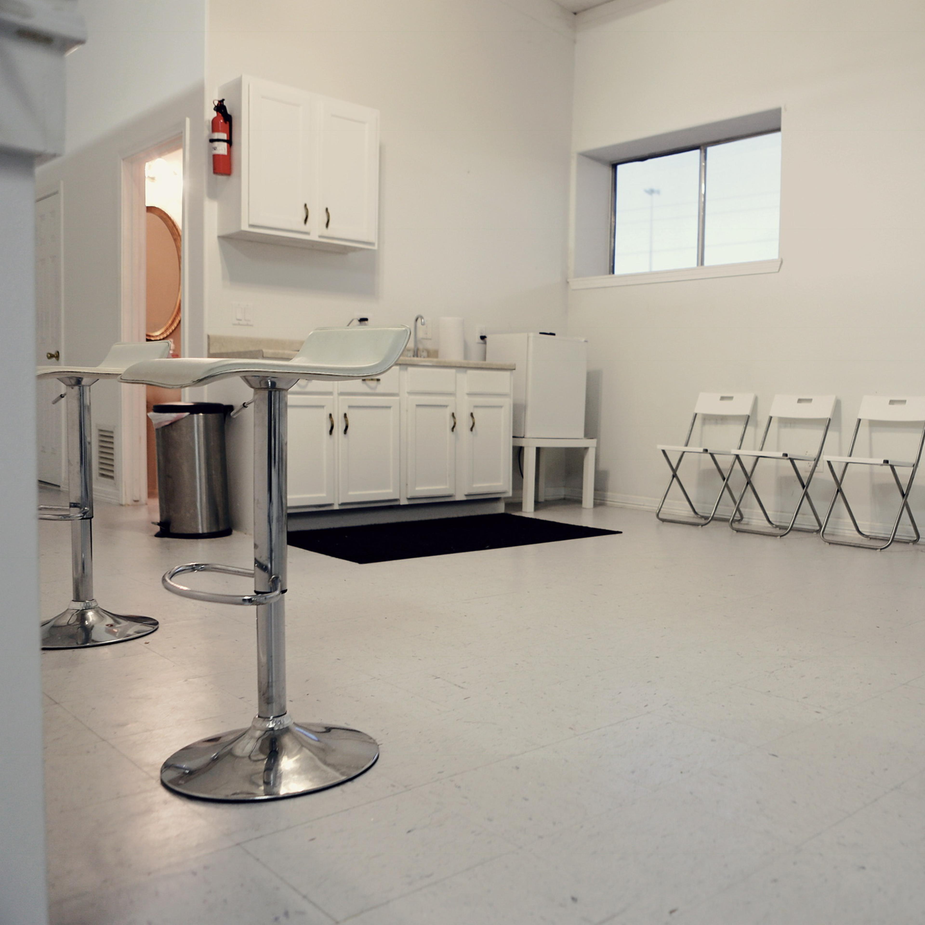 A minimalist waiting room with a row of white folding chairs, a kitchenette area, and a large window letting in natural light.