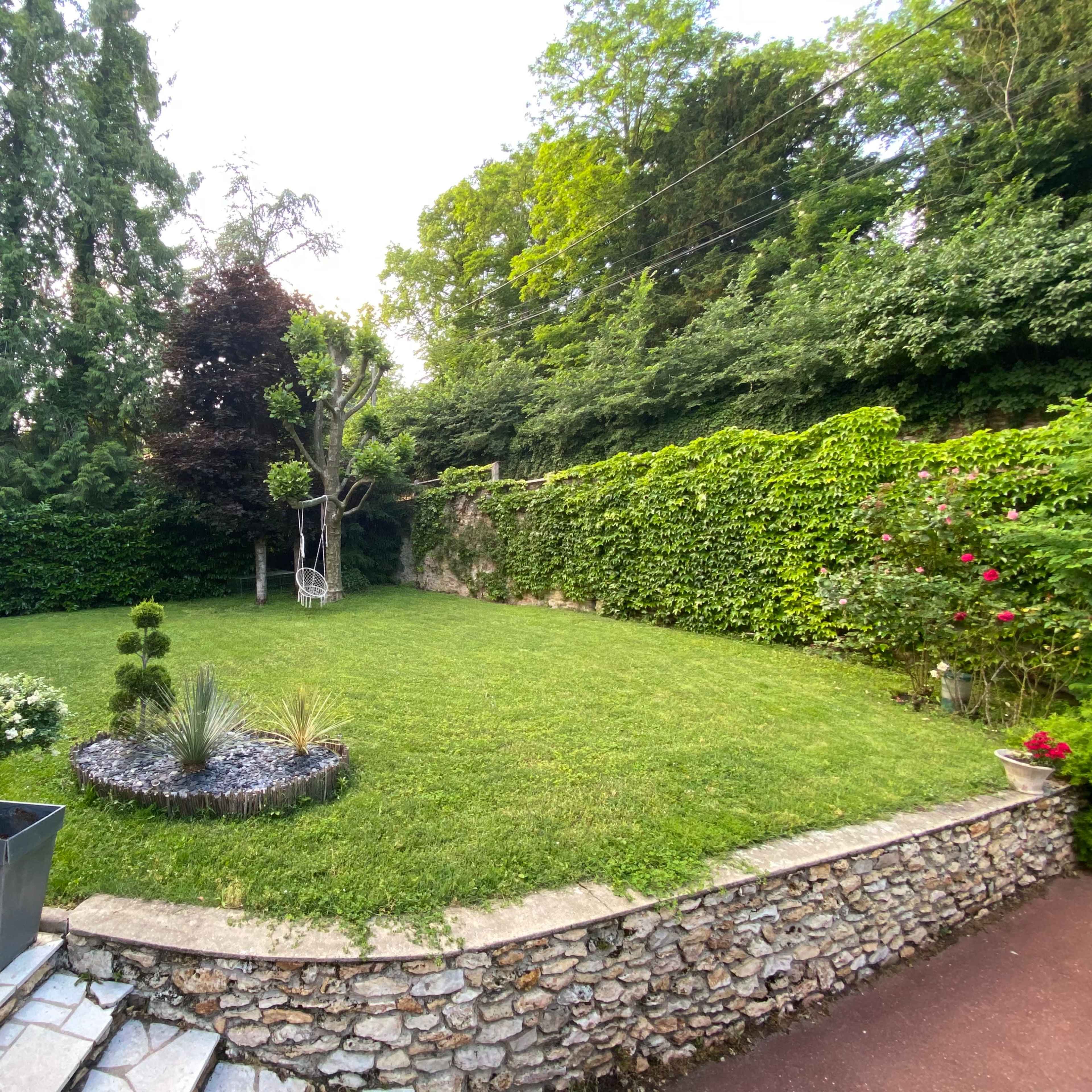 A well-maintained garden with a gravel border, surrounded by lush greenery and trees, featuring a small lawn, plants, and a decorative chair.