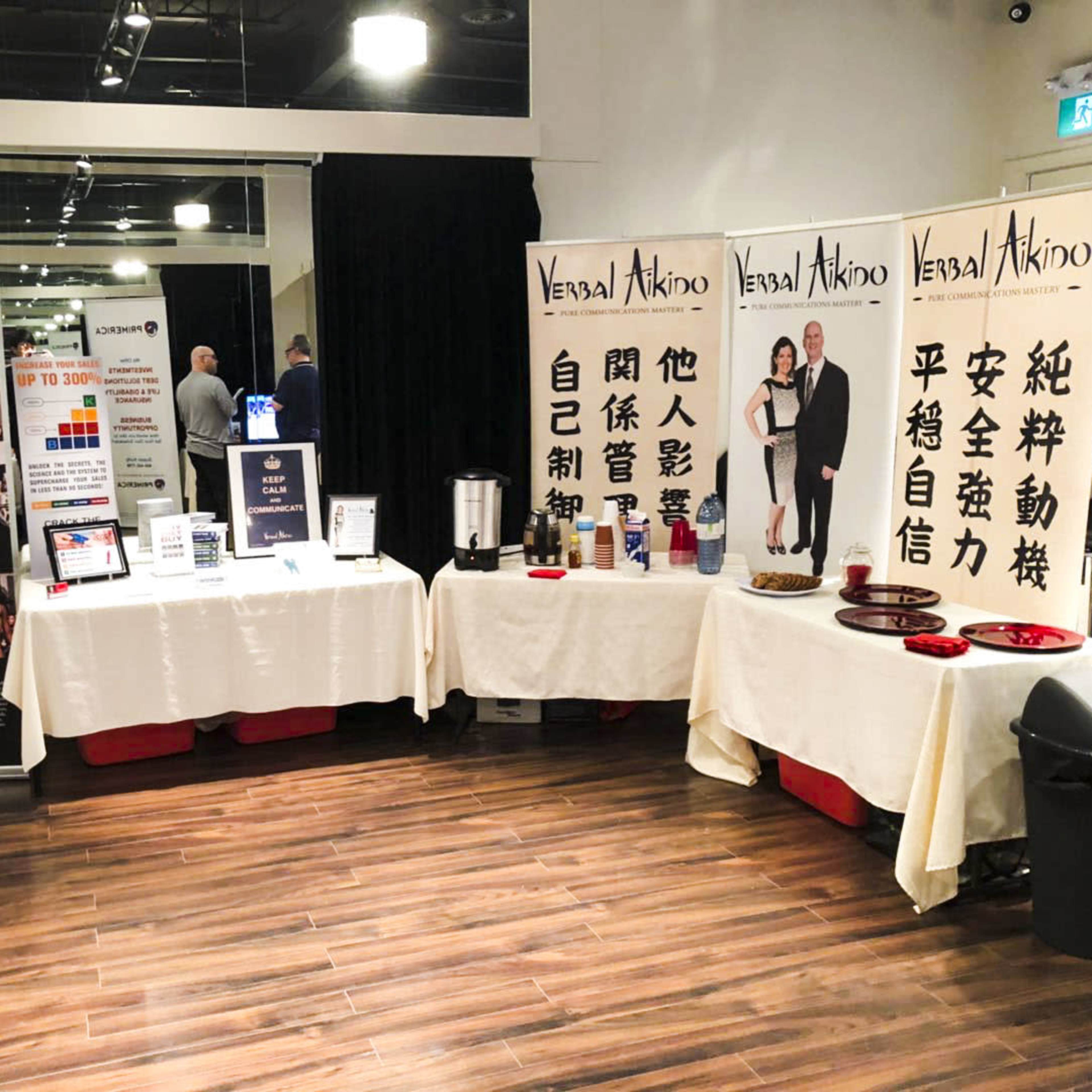 A display area with several tables covered in cloth, featuring promotional banners and informational materials for a community event related to martial arts or self-defense.