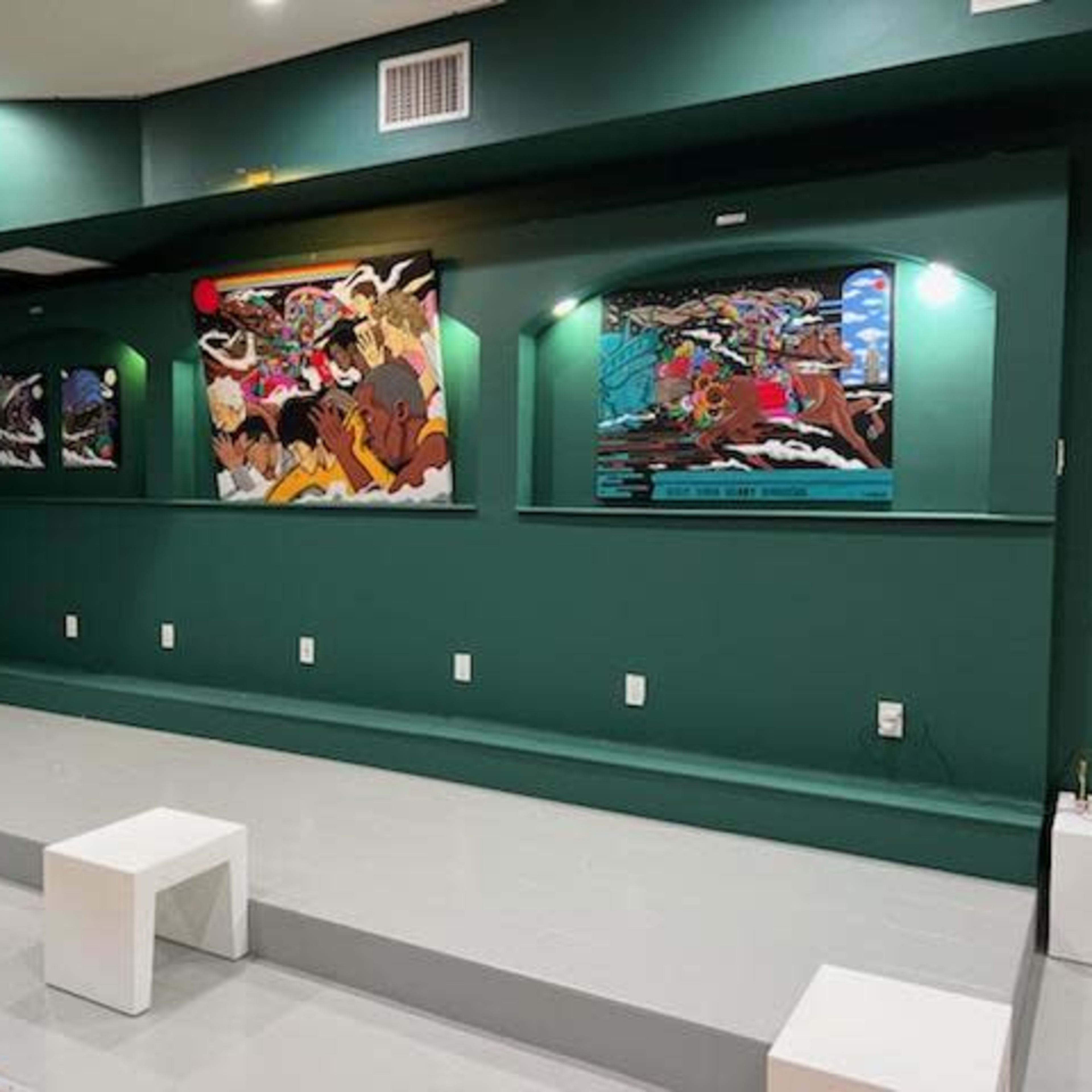 A green-walled gallery space with framed artwork displayed on the walls and white benches arranged on a gray floor.