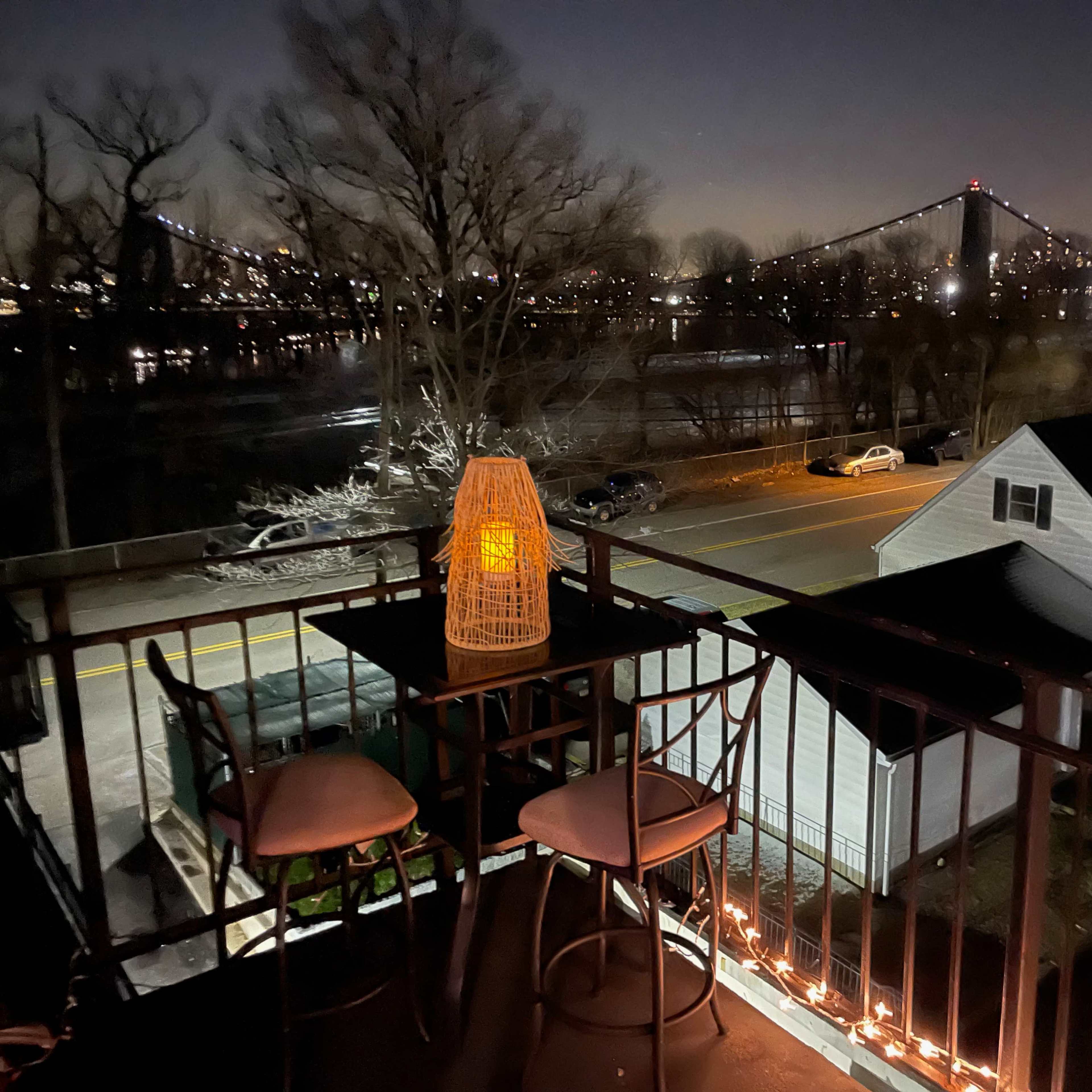 A rooftop balcony with two bar stools and a lantern, overlooking a river and a bridge illuminated against a night sky.